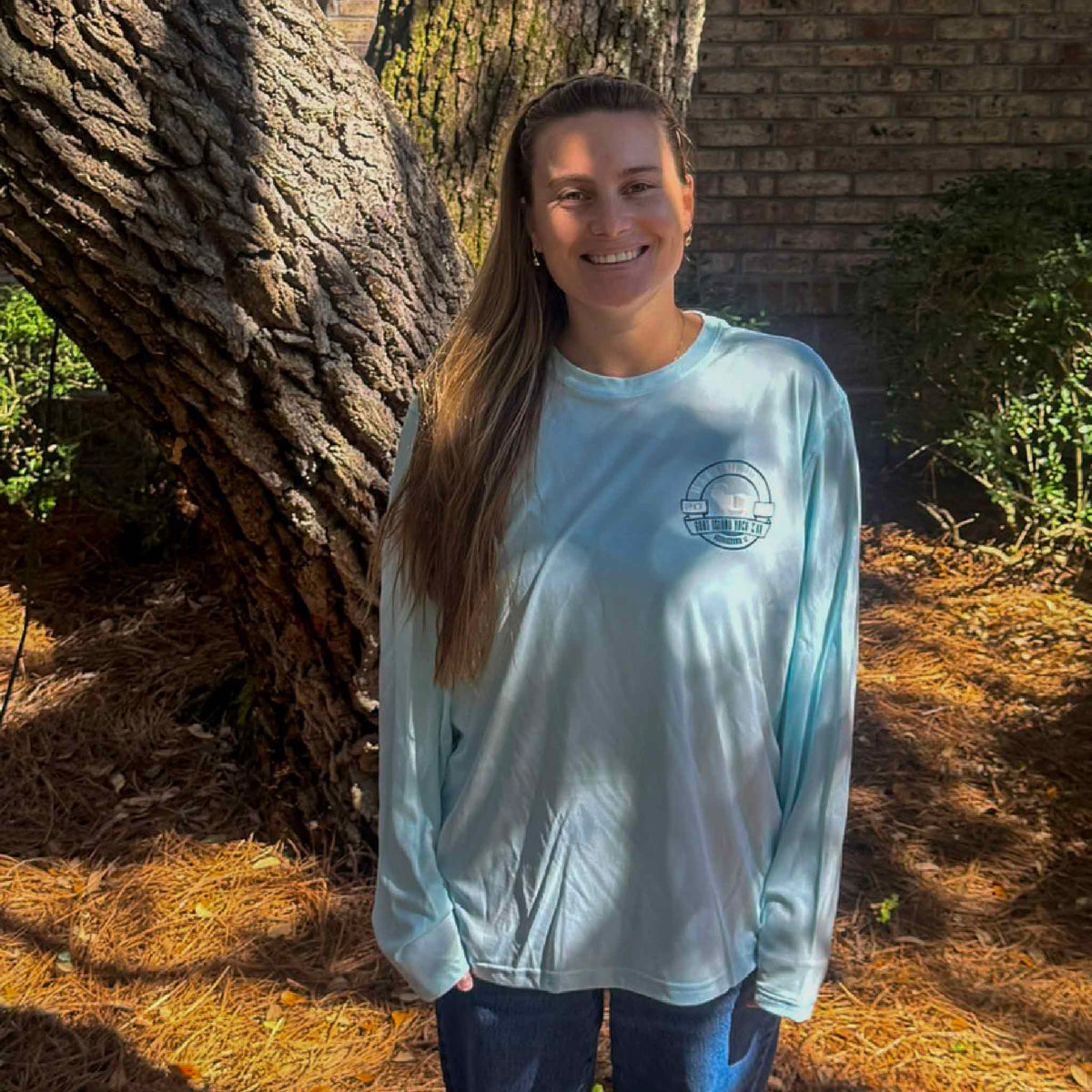 Kyra modeling the front of the Goat Island Yacht Club light blue long sleeve performance fishing shirt outdoors in Pawleys Island, South Carolina. The lightweight polyester crewneck features a small Georgetown SC yacht club logo on the left chest, long sleeves, and a relaxed silhouette. Coastal apparel available at Gray Man Gallery.