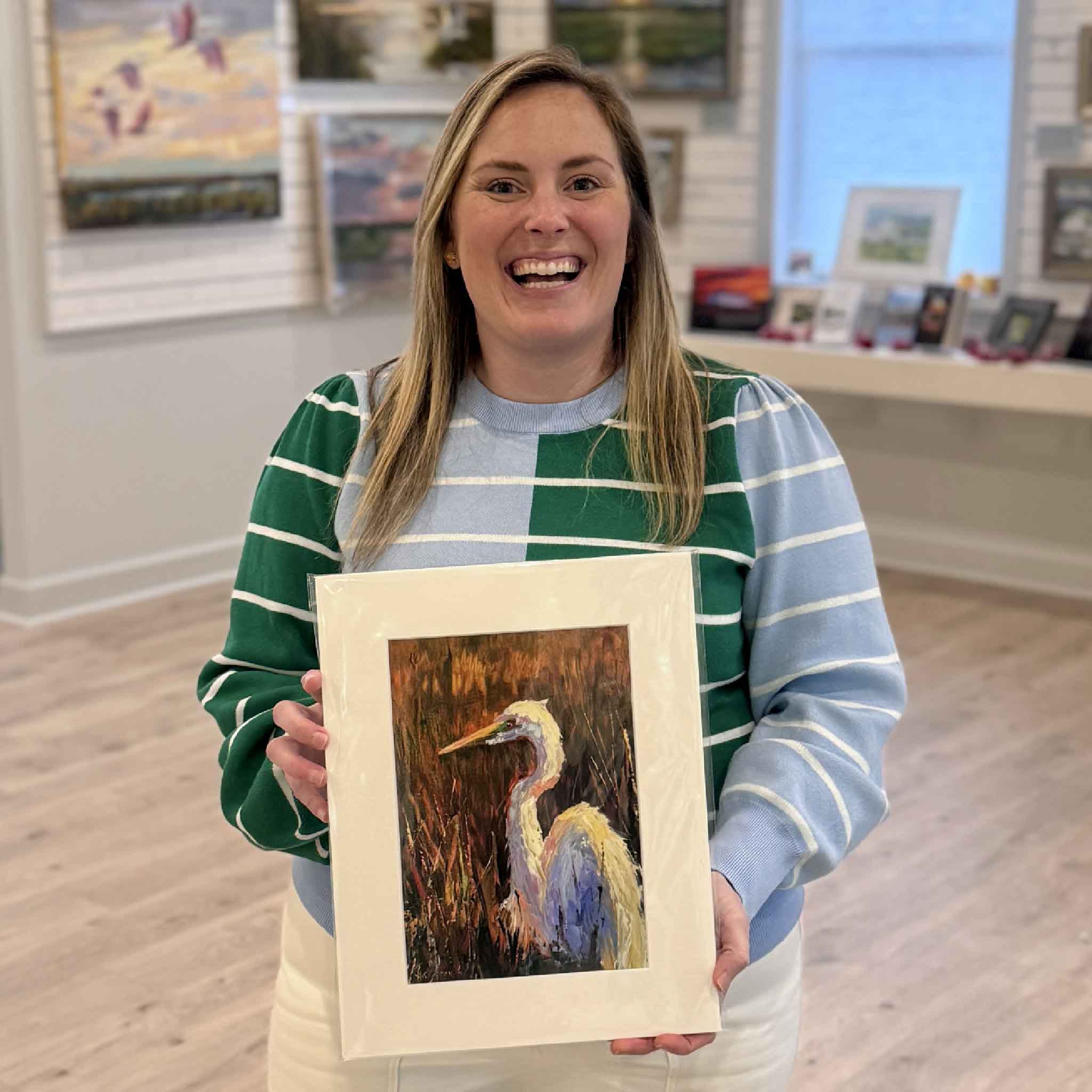 Gray Man Gallery team member holding Golden Marsh Light giclée print by Jeanne O’Neal featuring a white egret in marsh grasses, photographed inside Pawleys Island art gallery, showing scale and presentation of Lowcountry coastal artwork and local South Carolina artist prints