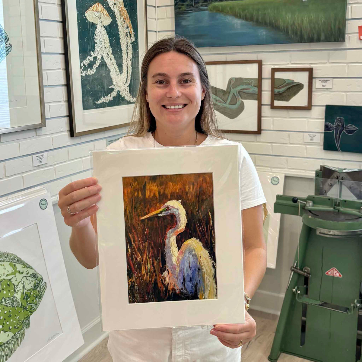 Gray Man Gallery team member holding Golden Marsh Light giclée print by Jeanne O’Neal featuring a white egret in marsh grasses, photographed inside Pawleys Island art gallery, showing scale and presentation of Lowcountry coastal artwork and local South Carolina artist prints