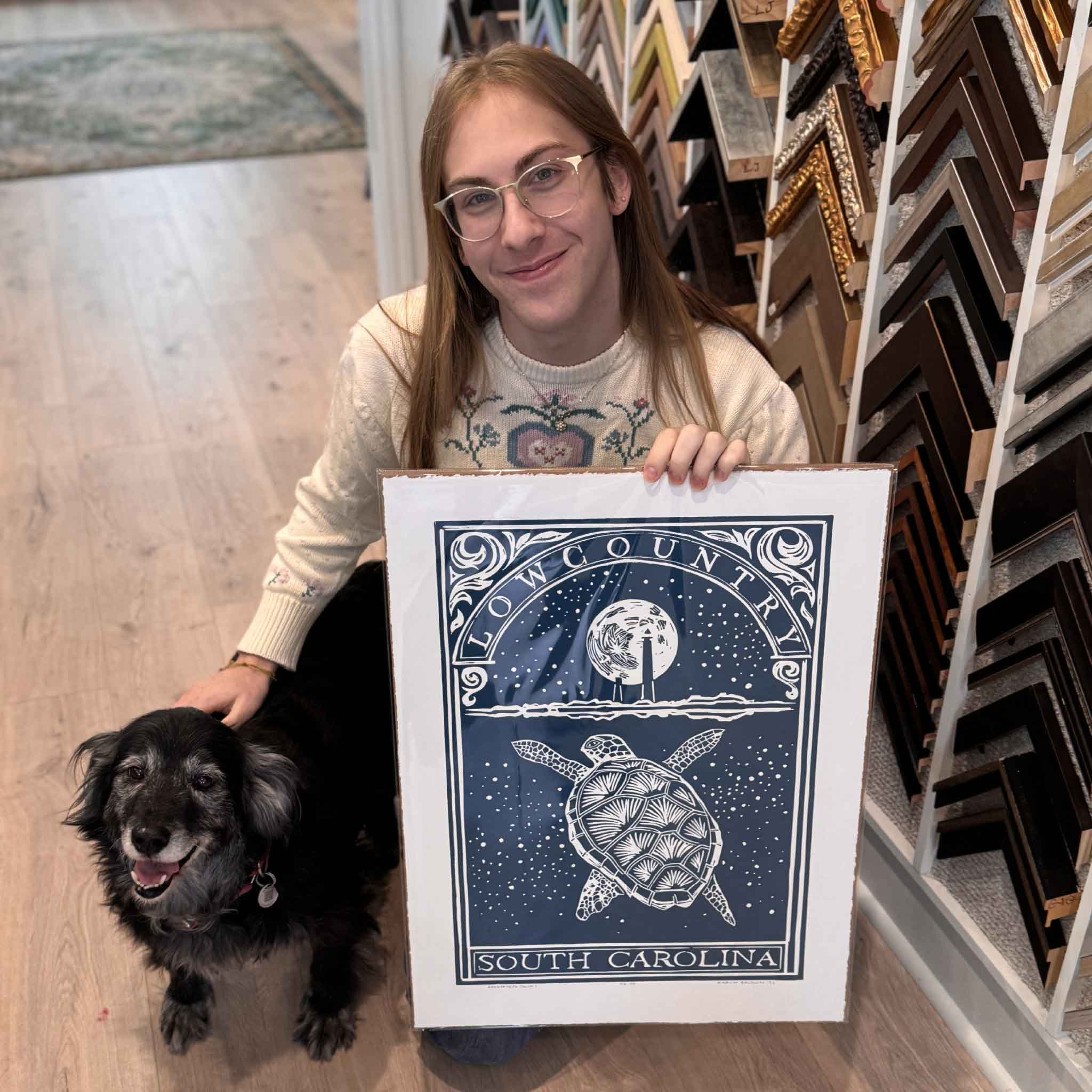 Gray Man Gallery team member holding Buddy, our rescue dog, and Loggerhead, an original linocut by Aaron Baldwin, shown for scale inside the gallery.
