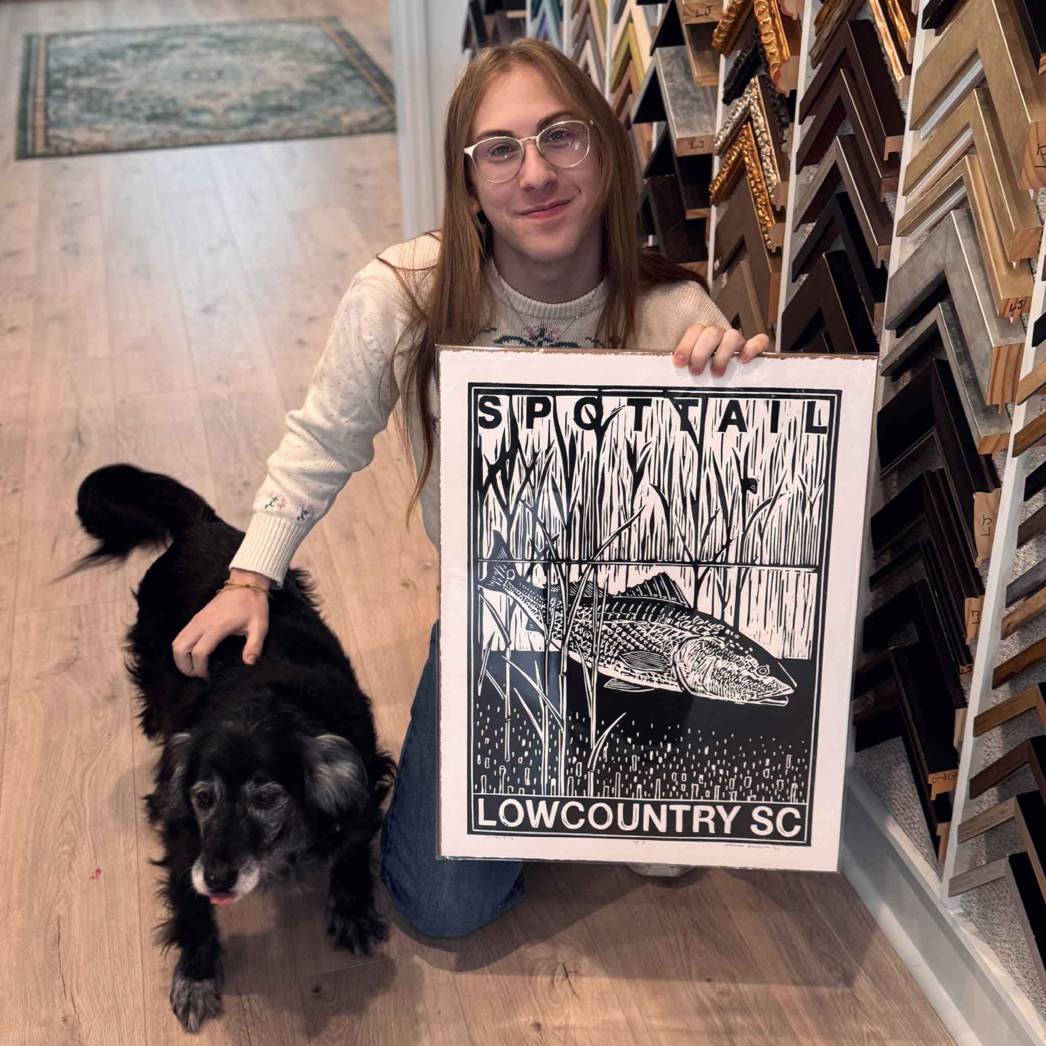 Gray Man Gallery team member holding Spottail II by Aaron Baldwin, an original linocut inspired by the Lowcountry marsh and coastal wildlife, AND Buddy, our rescue pup.