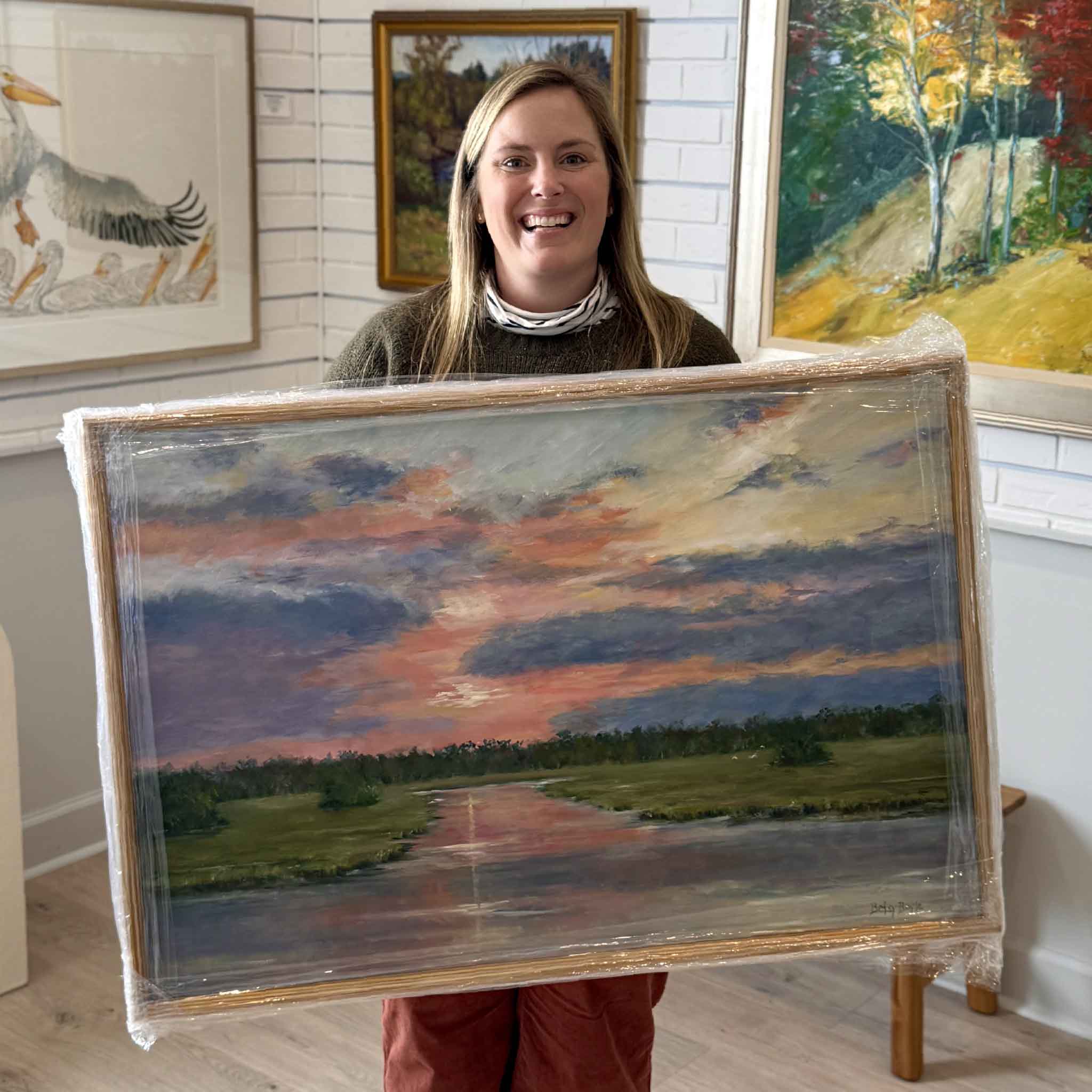 Bordy at Gray Man Gallery in Pawleys Island holding “Sweet Surrender,” a 36x24 original oil painting by local Lowcountry artist Betsy Boyle, featuring a colorful coastal marsh sunset scene professionally framed by the gallery