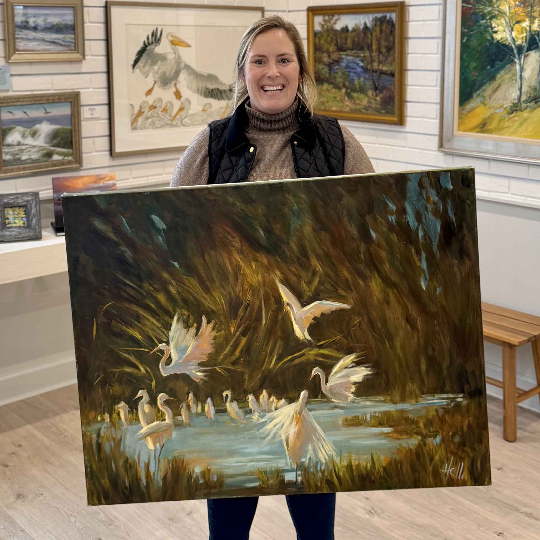Gray Man Gallery team member holding “Lowcountry Spa” original oil painting by Helli Luck featuring egrets in a Lowcountry marsh in Pawleys Island, South Carolina
