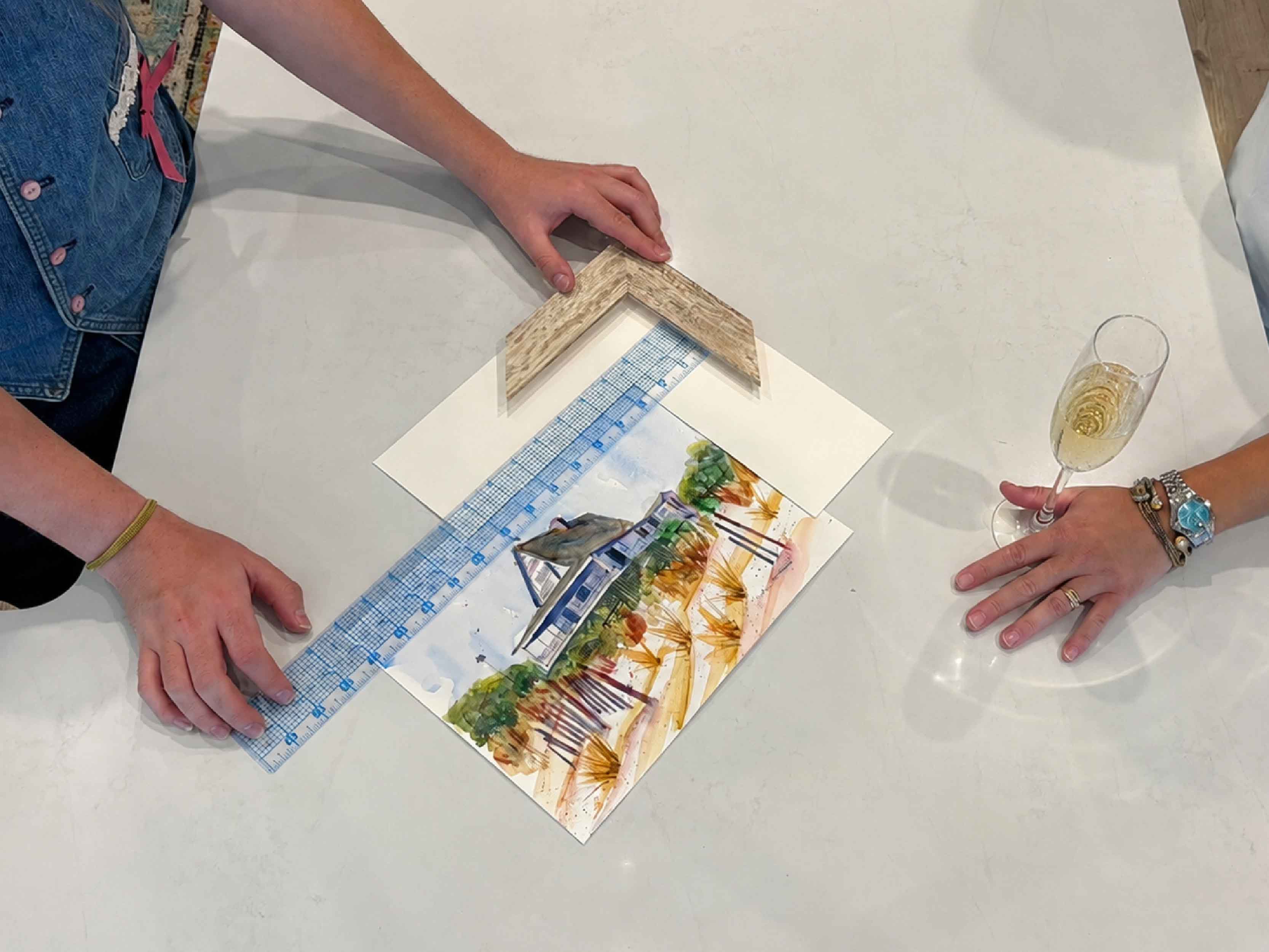 Overhead view of a custom framing consultation at Gray Man Gallery in Pawleys Island, South Carolina, where Kyra measures original watercolor artwork, compares custom wood frame moulding, discusses matting and proportions, and designs a conservation-quality custom picture frame with the client.