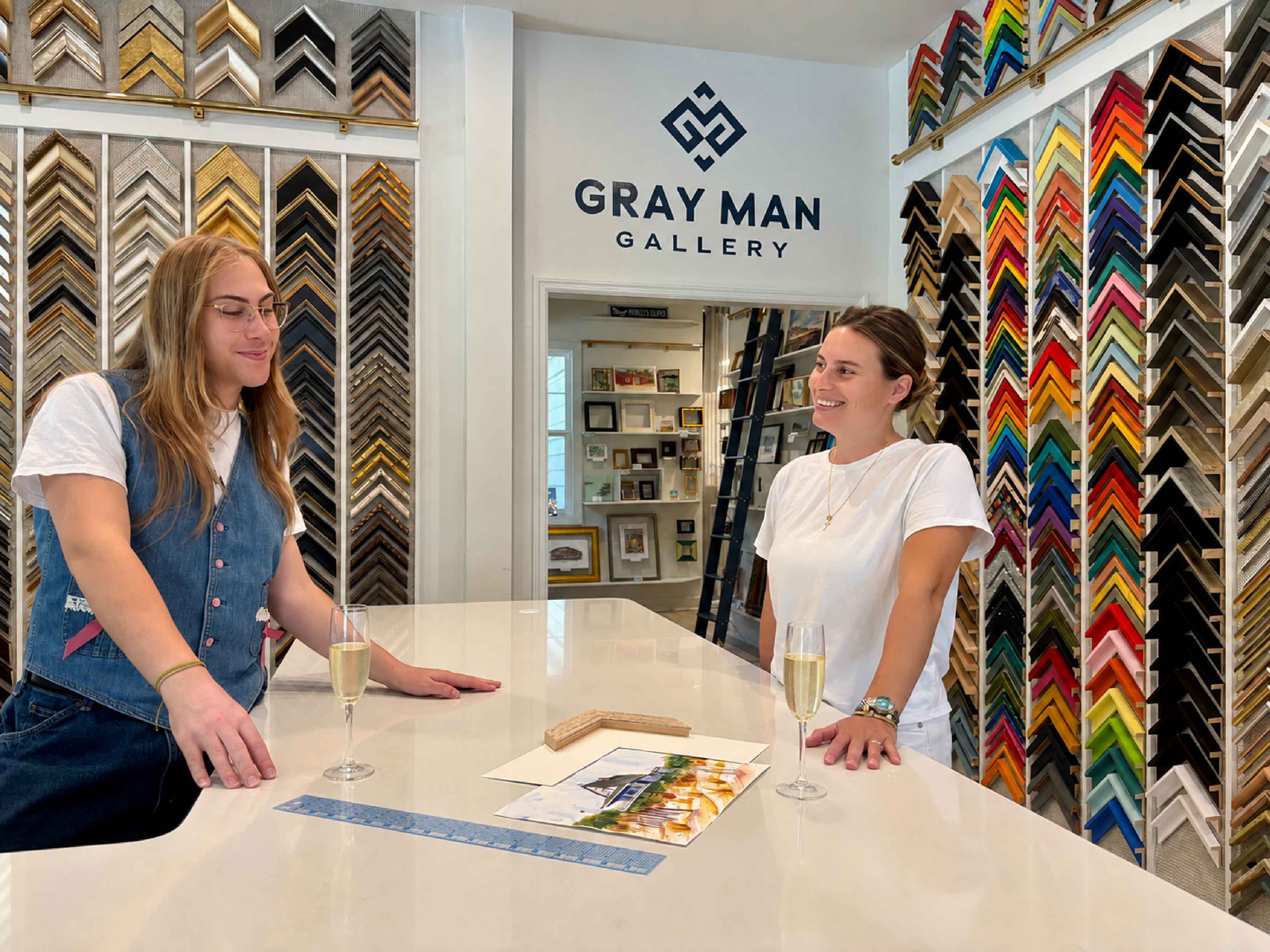 Professional custom framing consultation at Gray Man Gallery in Pawleys Island, South Carolina, where Kyra and Alex help a customer select the perfect frame and mat for original artwork using hundreds of custom moulding samples, museum-quality materials, and expert conservation framing advice.