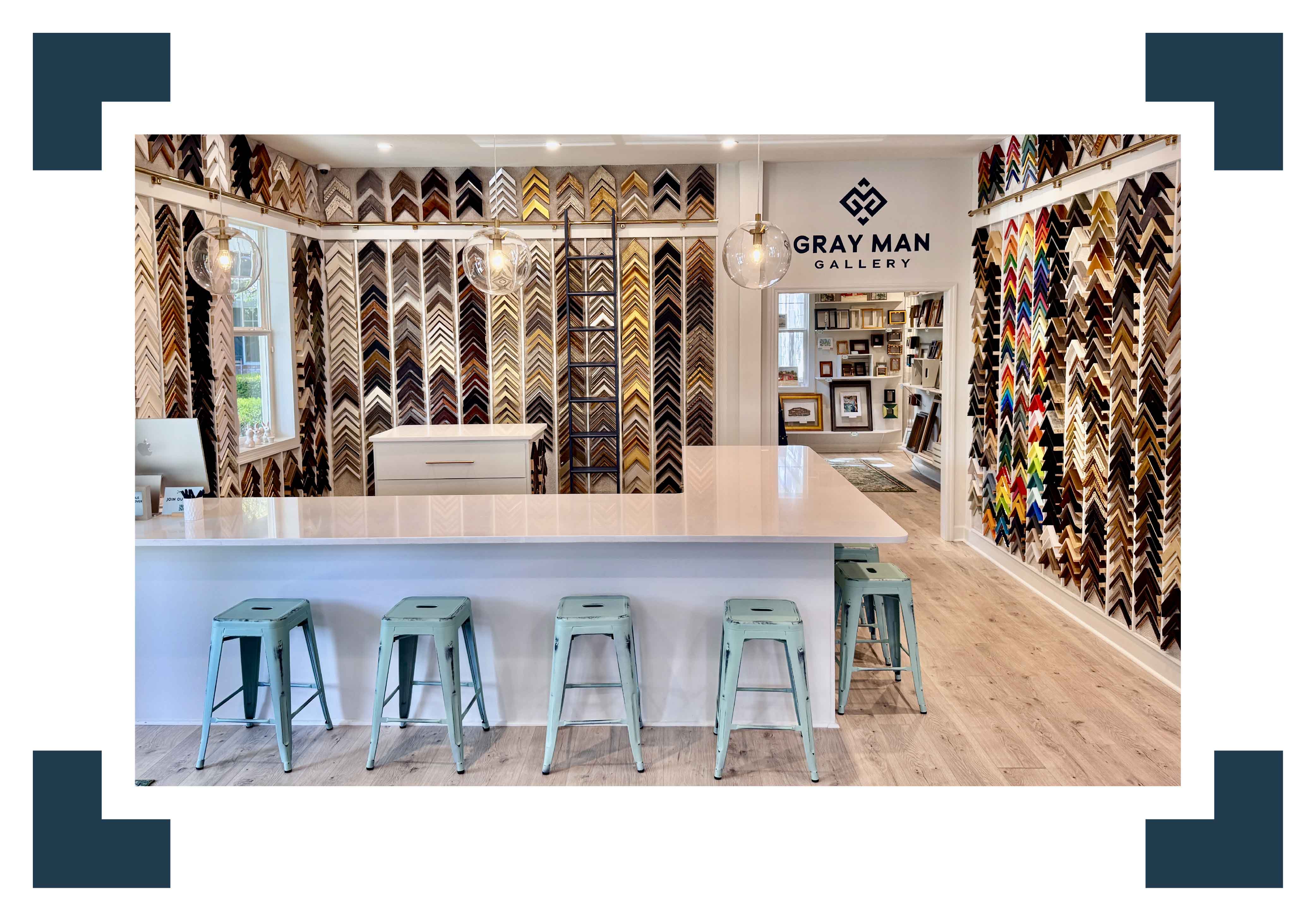 Custom framing design bar at Gray Man Gallery in Pawleys Island, South Carolina, featuring a bright, welcoming workspace with a large white consultation table, aqua stools, and walls filled with frame corner samples in a wide range of finishes and styles, showcasing the gallery’s extensive custom framing options and boutique in-house craftsmanship in a clean, coastal Lowcountry setting