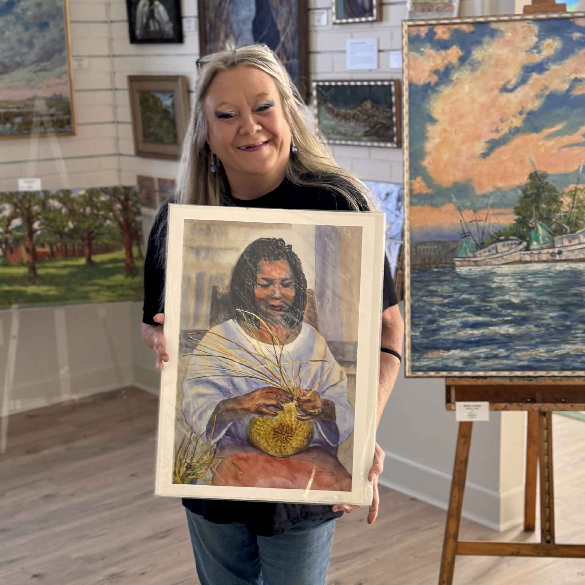 Charlotte holding a watercolor print titled Basket Lady by Gail Joley depicting a Gullah Geechee woman weaving a traditional sweetgrass basket, celebrating Lowcountry craftsmanship and cultural heritage. Pictured at Gray Man Gallery.