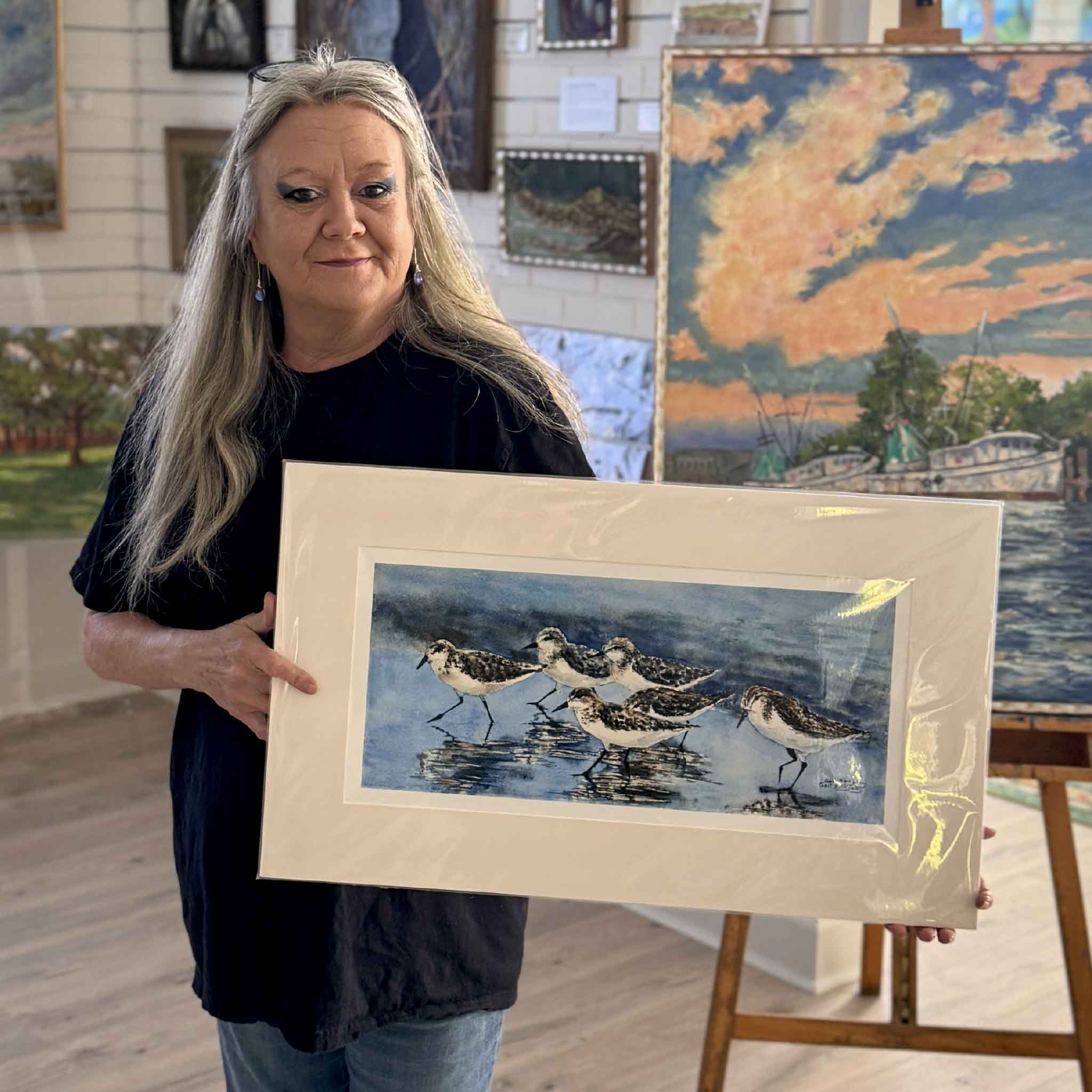 Charlotte holding the Sanderlings watercolor print by Gail Joley showing a small flock of shorebirds walking along wet sand, reflected in shallow coastal water, photographed at Gray Man Gallery in Pawleys Island.