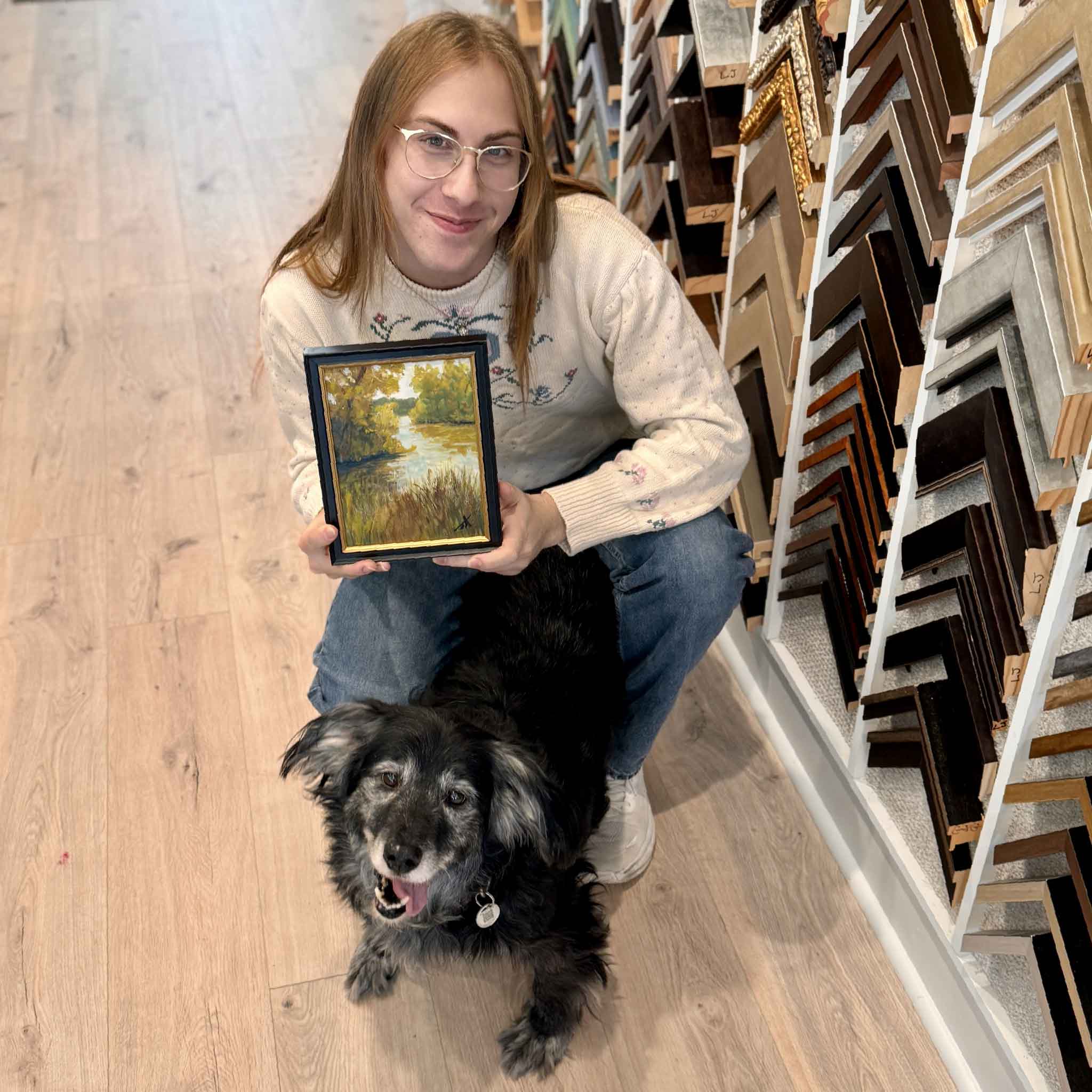 Jamie Arnold’s original oil Along the Quiet Water shown framed and held in the gallery for scale, highlighting its warm light, serene water, and Lowcountry landscape details. And our rescue dog, Buddy.