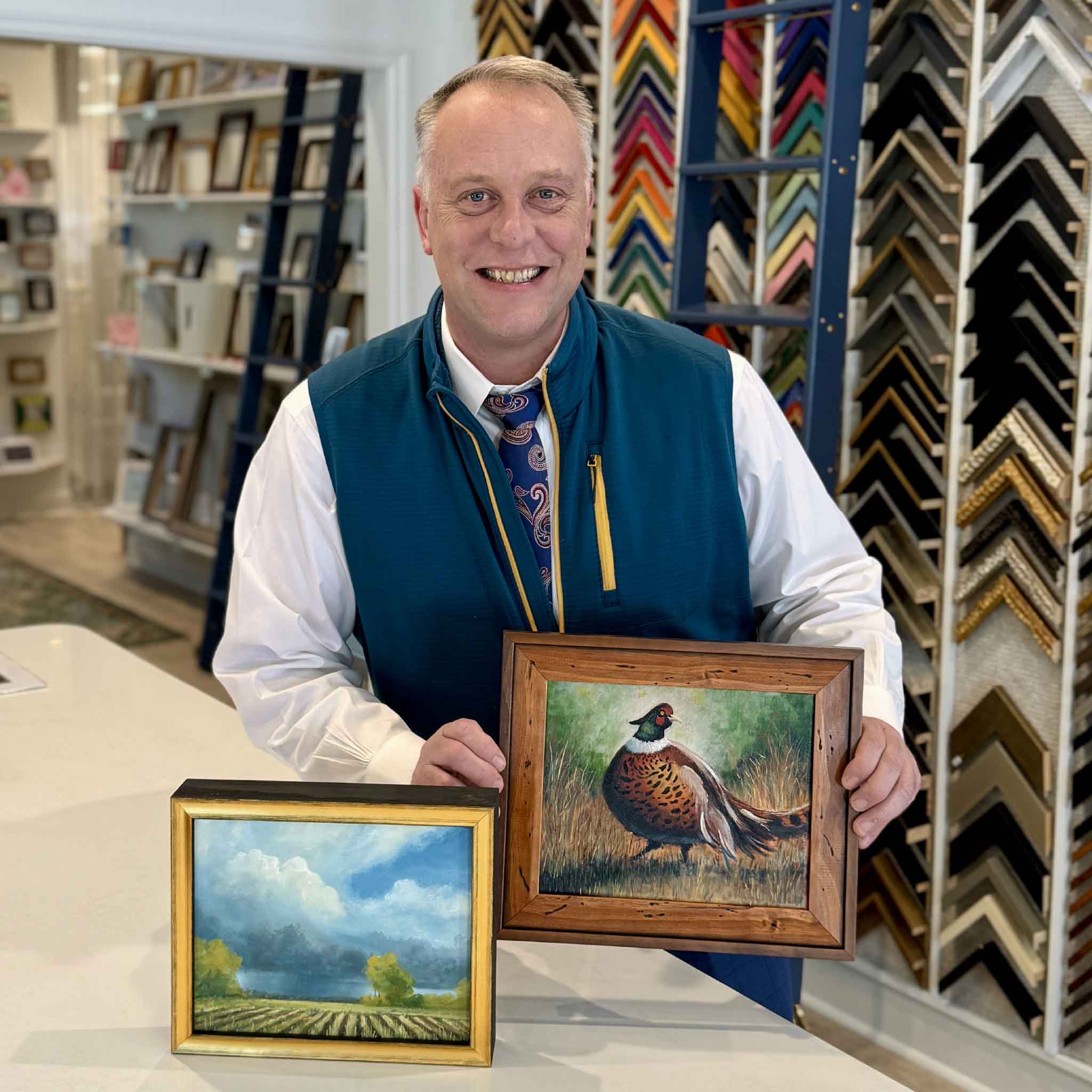 Jamie Arnold holding two original oil paintings at Gray Man Gallery in Pawleys Island, South Carolina, featuring Birds of a Pheasant wildlife artwork in a rustic wood frame and Mayfields Lowcountry landscape in a gold frame, showcasing fine local art and custom framing