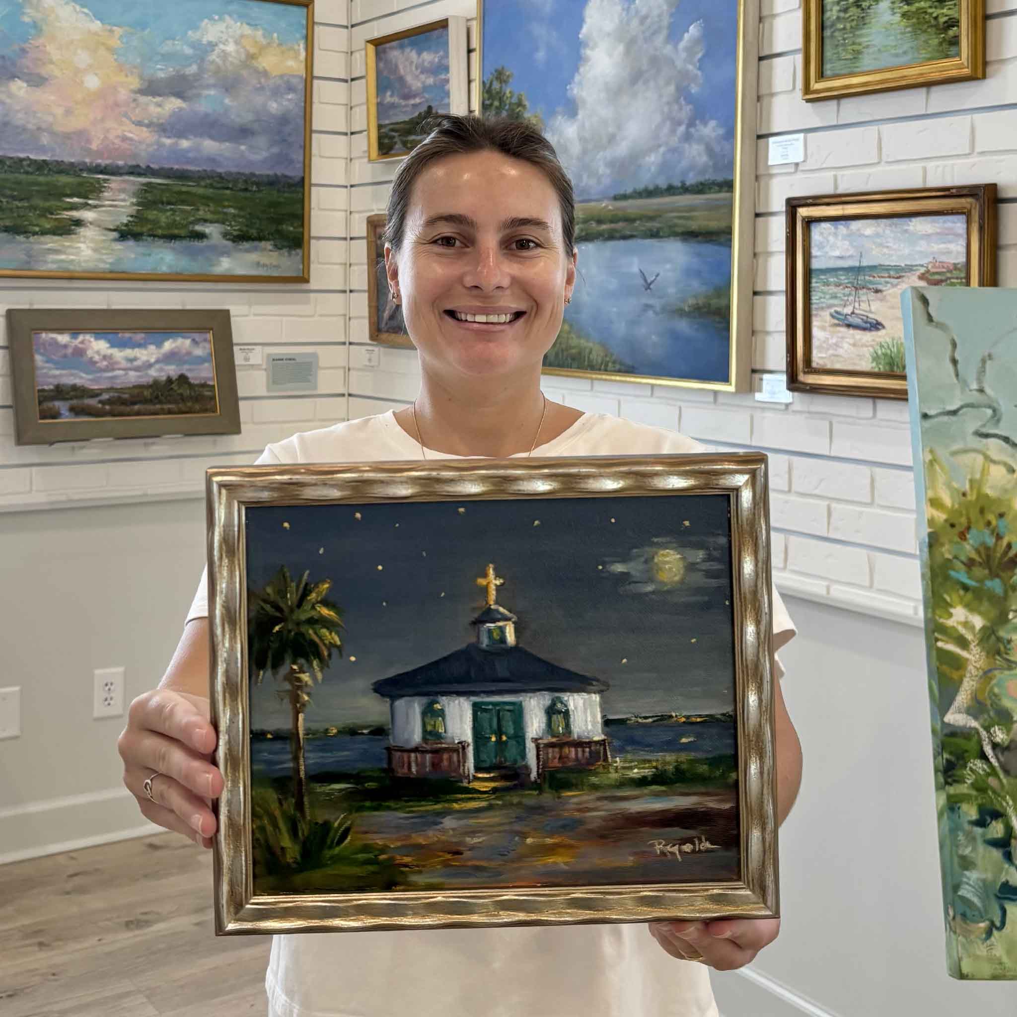 Kyra holds Moonlight Over the Chapel by Jenny Reynolds inside Gray Man Gallery in Pawleys Island, South Carolina. This original oil painting depicts a moonlit coastal chapel with glowing windows, a full moon reflected on the water, and a tranquil Lowcountry evening, beautifully presented in Jenny Reynolds' signature champagne frame.