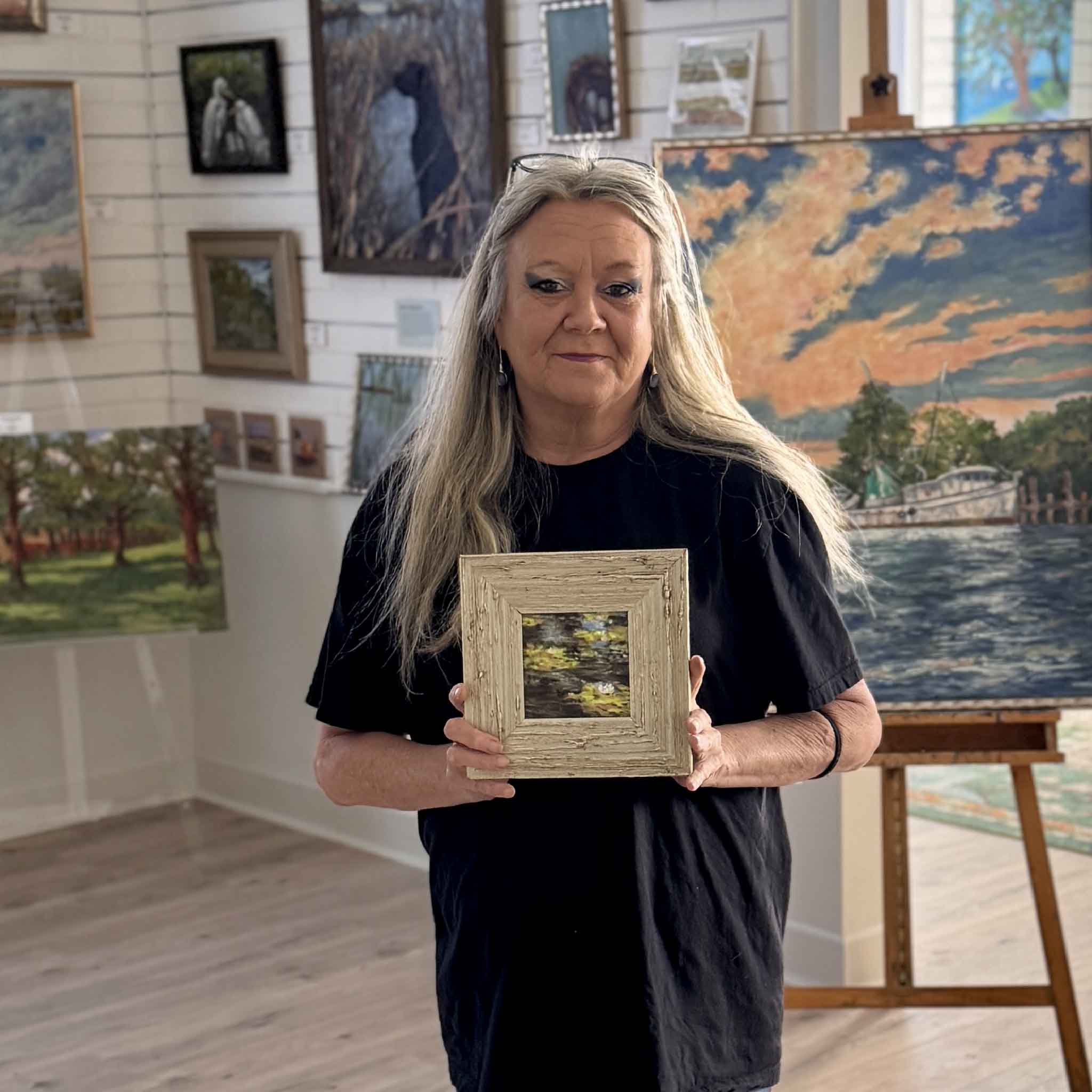 Charlotte holding Joanne Willoughby's Lilies I, a small original 4x4 pastel artwork framed and ready to display, shown for scale inside Gray Man Gallery among original Lowcountry paintings and coastal art.