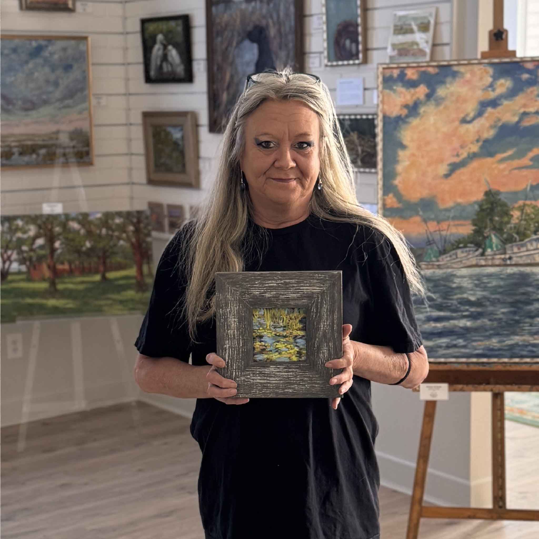 Charlotte holding Lowcountry artist Joanne Willoughby's original framed pastel Lilies III inside Gray Man Gallery, showcasing a small 4x4 water lily marsh scene with layered pastel texture and reflective coastal tones.