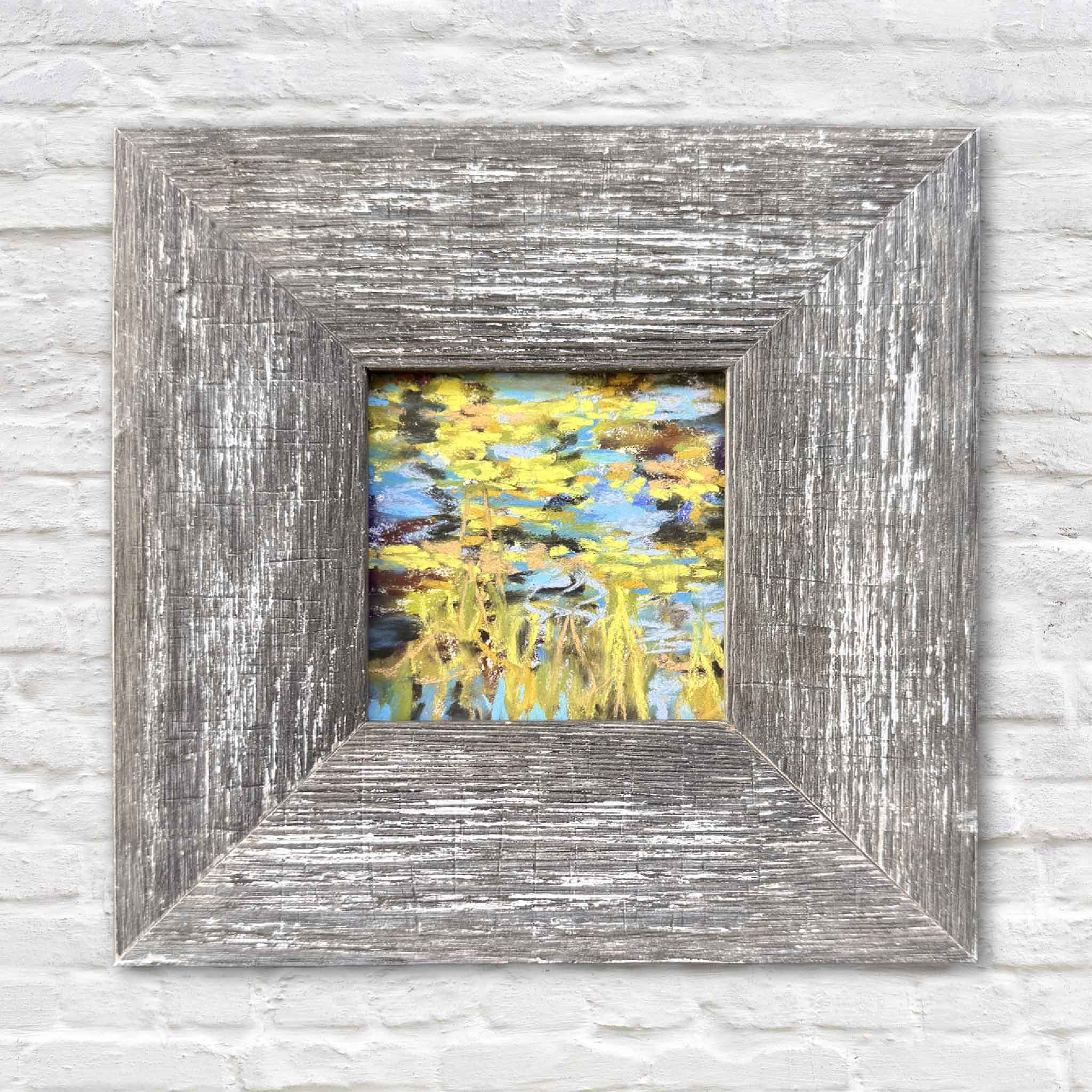 Original 4x4 pastel artwork Lilies III by Joanne Willoughby, featuring soft blue water, yellow-green lily pads, and marsh grasses, professionally framed in a rustic wood frame and displayed against a white brick wall at Gray Man Gallery.