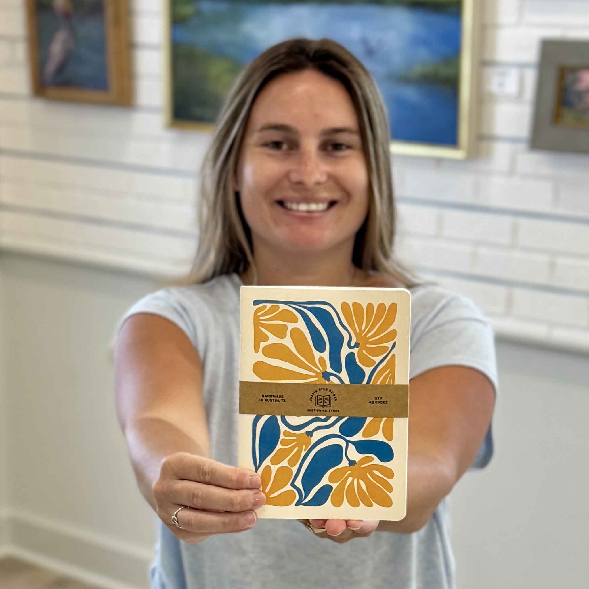 Kyra holding the Yellow Floral Notebook by Justin Ryan Books at Gray Man Gallery in Pawleys Island, South Carolina. This 5x7 handmade notebook features a cheerful boho-inspired floral cover and 48 blank pages for journaling, sketching, note-taking, and creative ideas.
