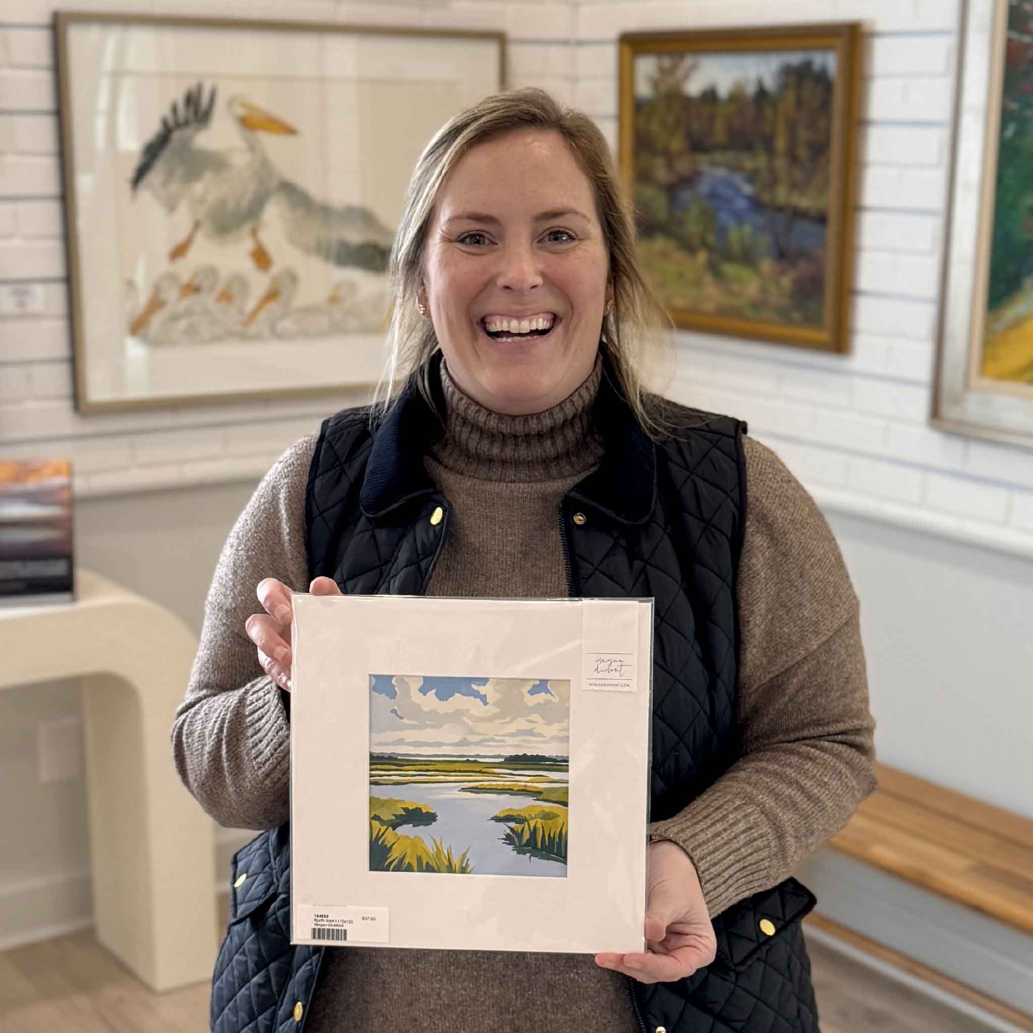 Smiling Gray Man Gallery staff member holding “North Inlet I,” an original 10x10 gouache marsh painting by local Lowcountry artist Megan DuMont, inside Gray Man Gallery in Pawleys Island, South Carolina, highlighting custom framing craftsmanship, coastal artwork, local artists, and boutique gallery interior.