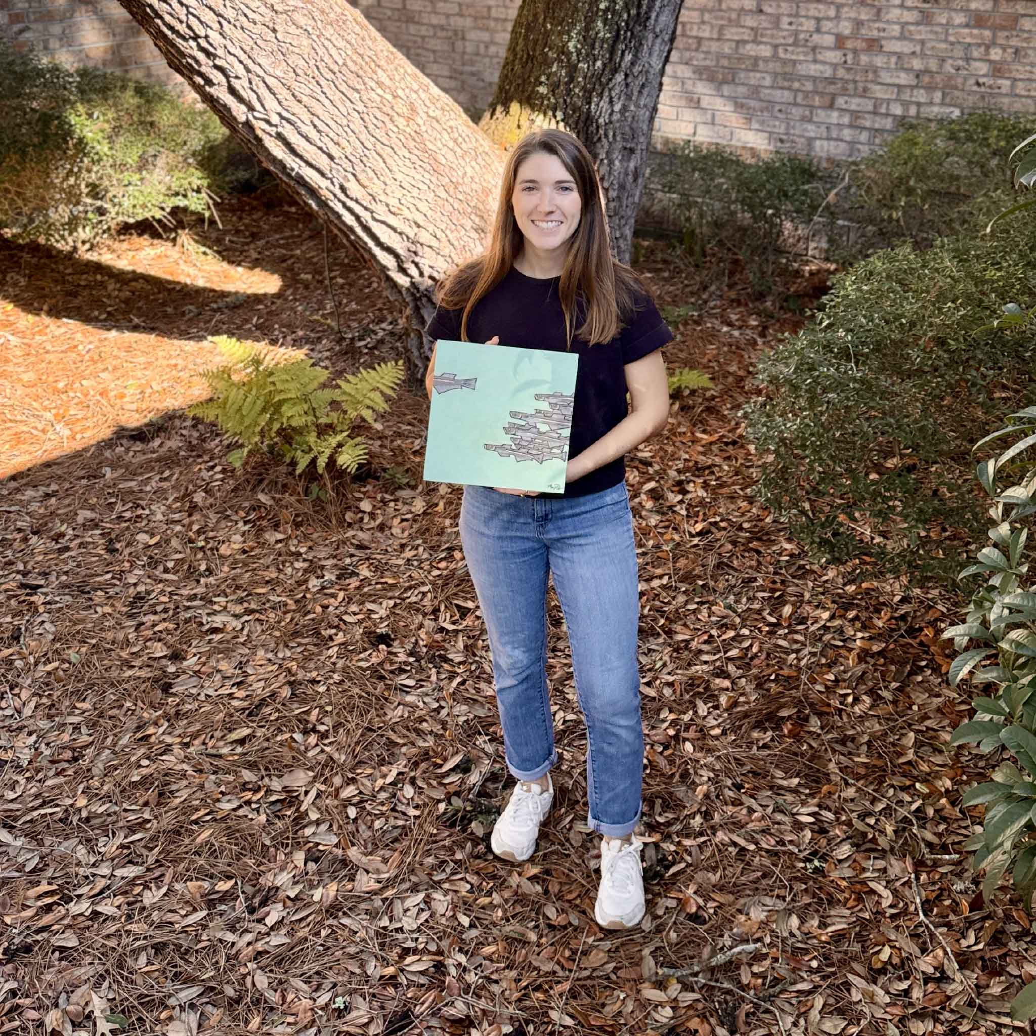 Artist Maggie Pelton holding her original 10 x 10 acrylic and charcoal painting Silverside outdoors, shown for scale and photographed in the courtyard at Gray Man Gallery.