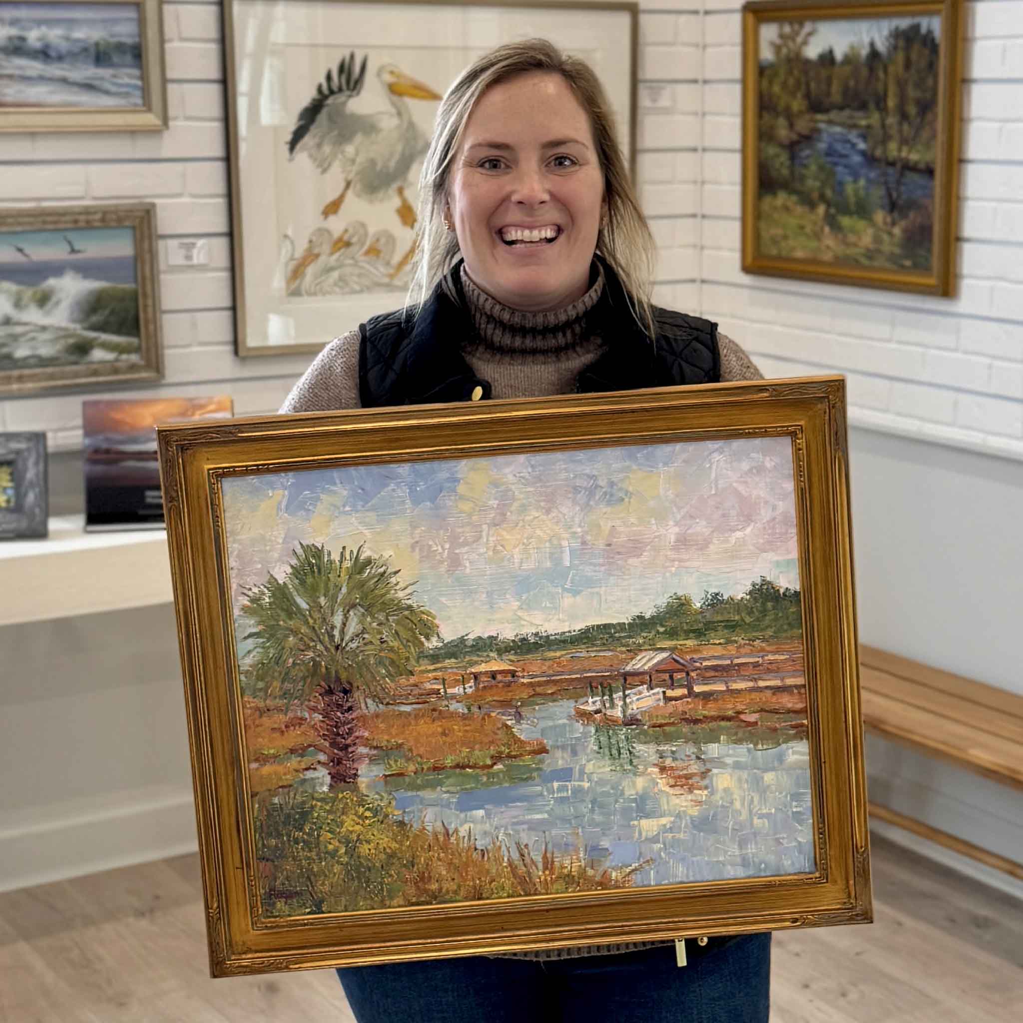 Gray Man Gallery staff member Bordy holding “Creek with Palm” by local artist Michael Rogan, an original oil painting of a Lowcountry marsh with palm tree and creek, photographed inside Gray Man Gallery in Pawleys Island, South Carolina
