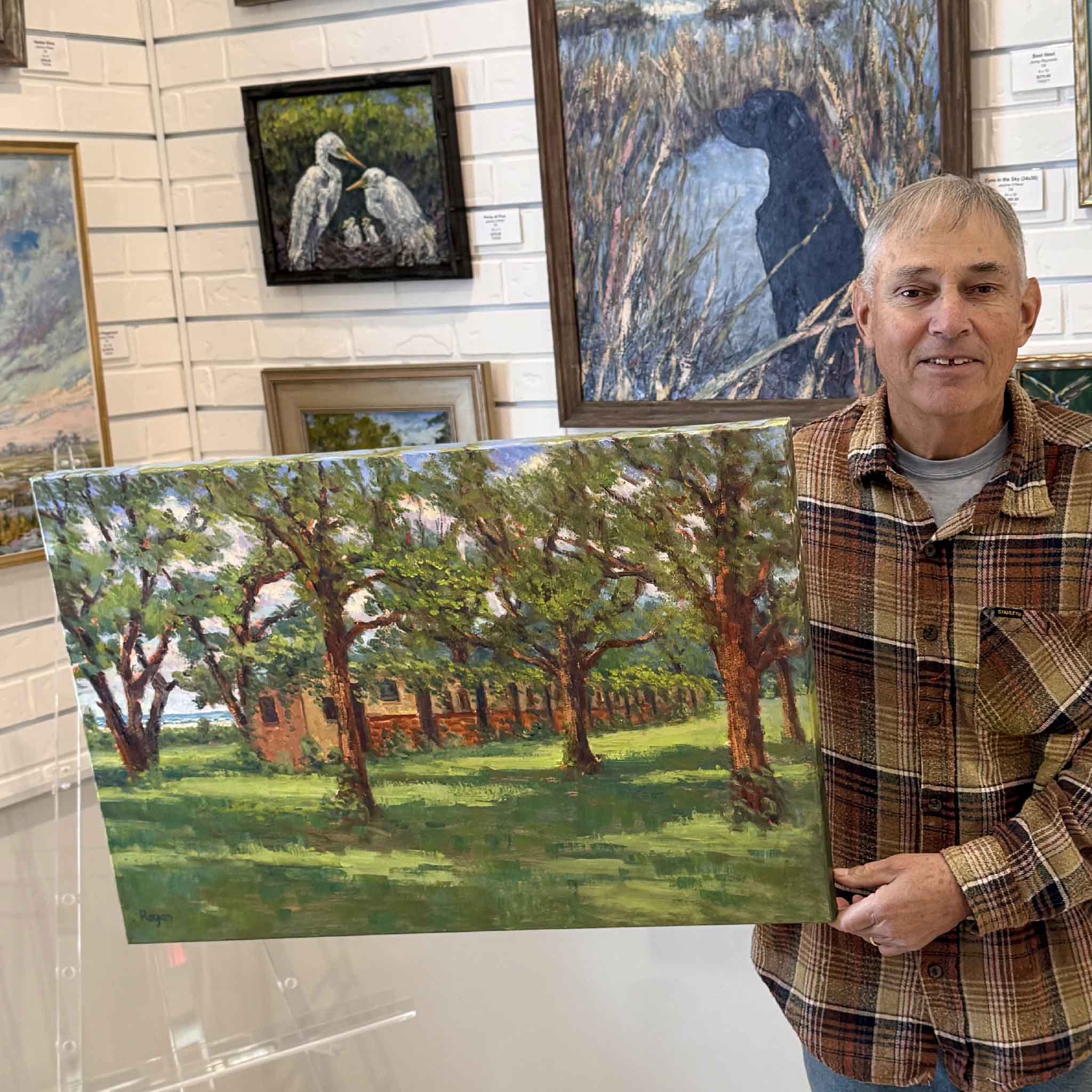 Local artist Michael Rogan holding his original gallery-wrapped oil painting, McKenzie Beach, depicting historic coastal cottages beneath live oaks with dappled sunlight and expressive brushwork. Pictured at Gray Man Gallery.