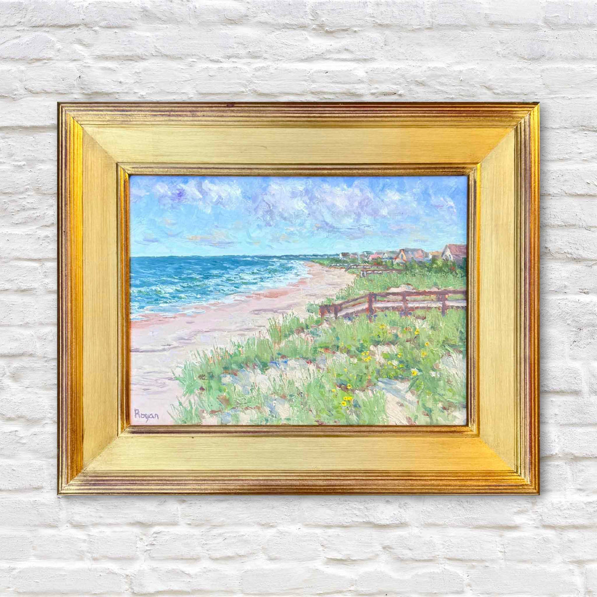 View to Inlet Point, a framed coastal landscape painting by local South Carolina artist Michael Rogan, depicting the shoreline near Inlet Point on Pawleys Island. Blue Atlantic Ocean waves meet a sandy beach bordered by grassy dunes, yellow wildflowers, rustic wooden fencing, and distant coastal cottages beneath a soft blue and lavender sky. Displayed in a wide traditional gold frame against the signature white brick wall at Gray Man Gallery in Pawleys Island, South Carolina.