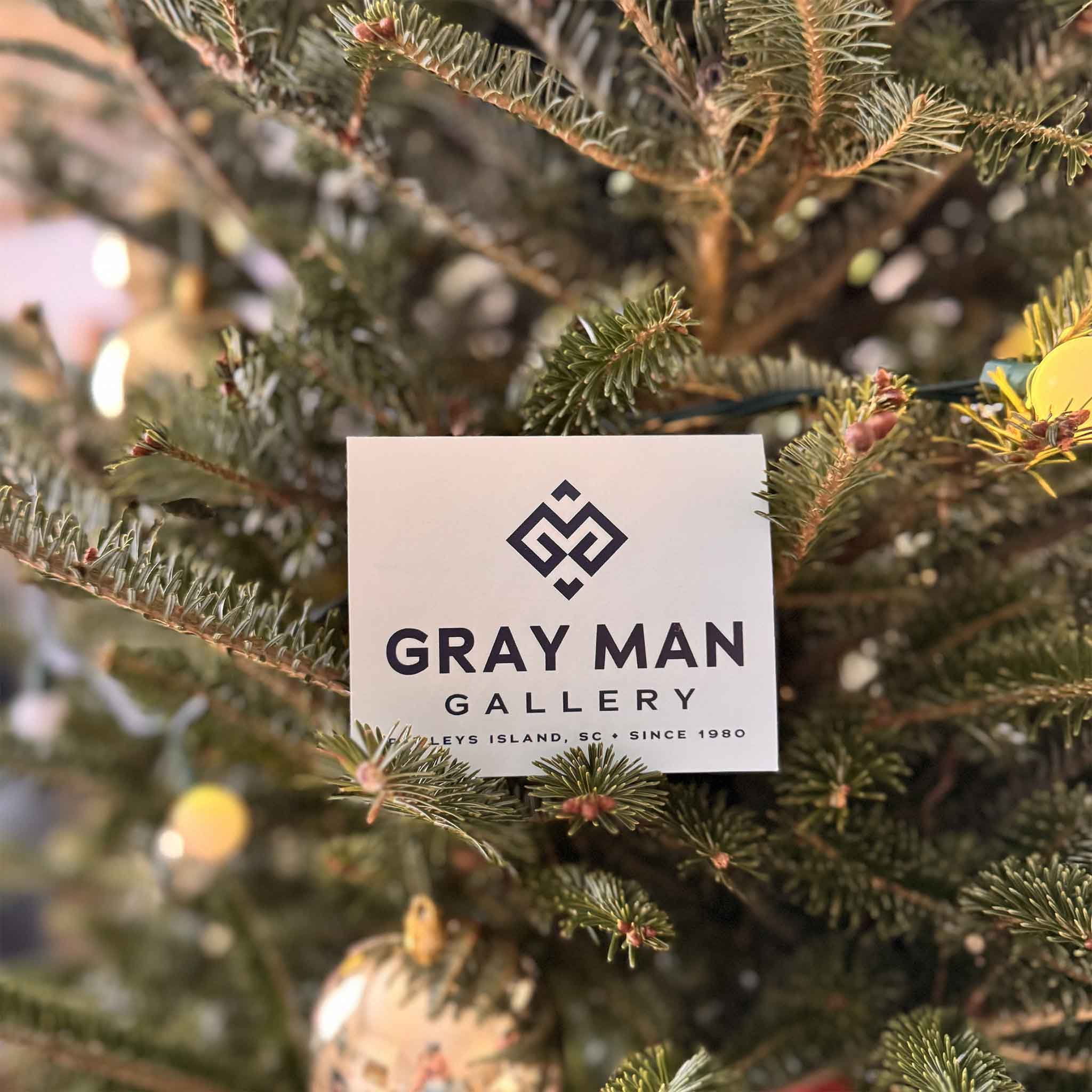 Gray Man Gallery physical gift card held in front of the Christmas tree inside the Pawleys Island art gallery.