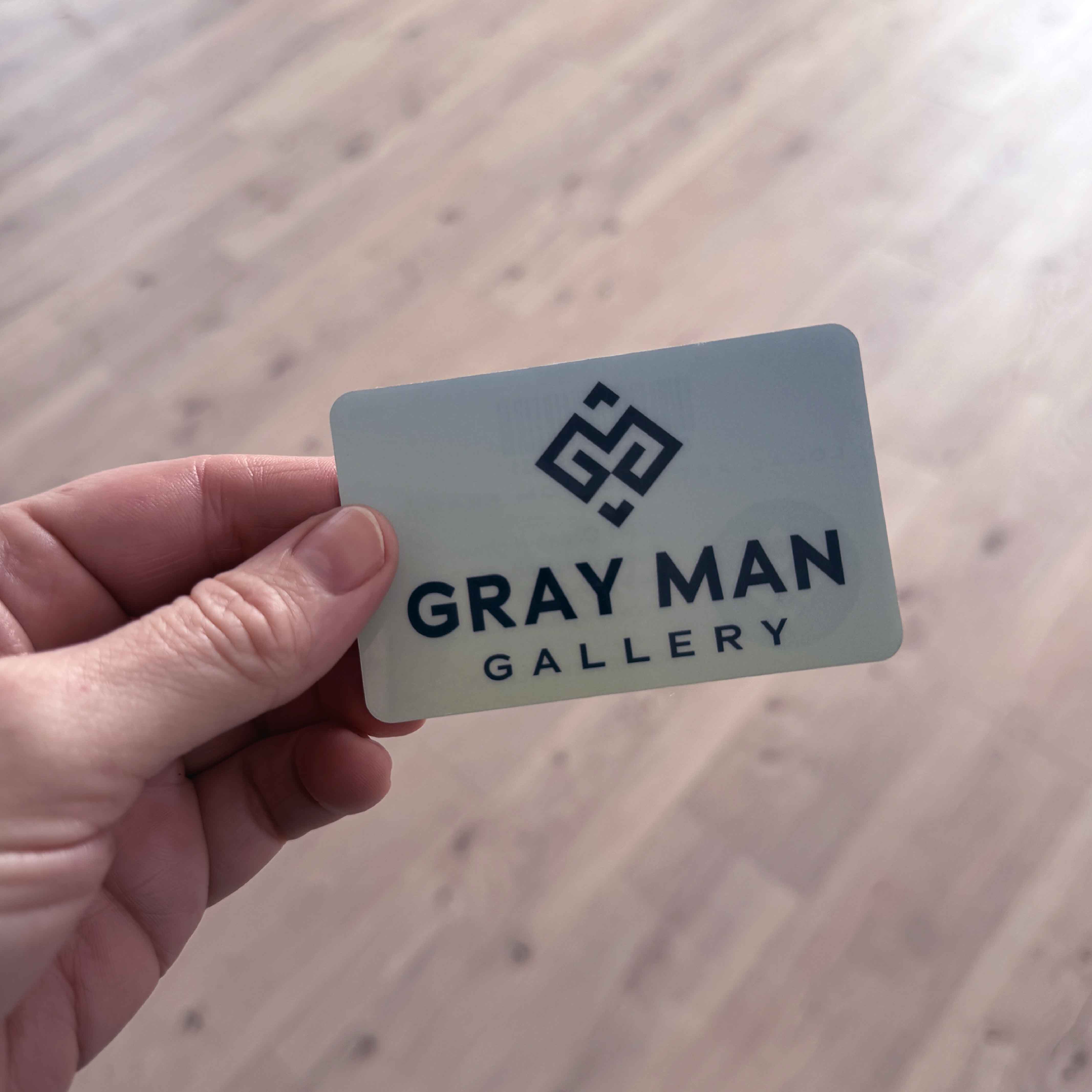 Hand holding a Gray Man Gallery physical gift card with coastal gradient logo design, photographed against a light wood background.
