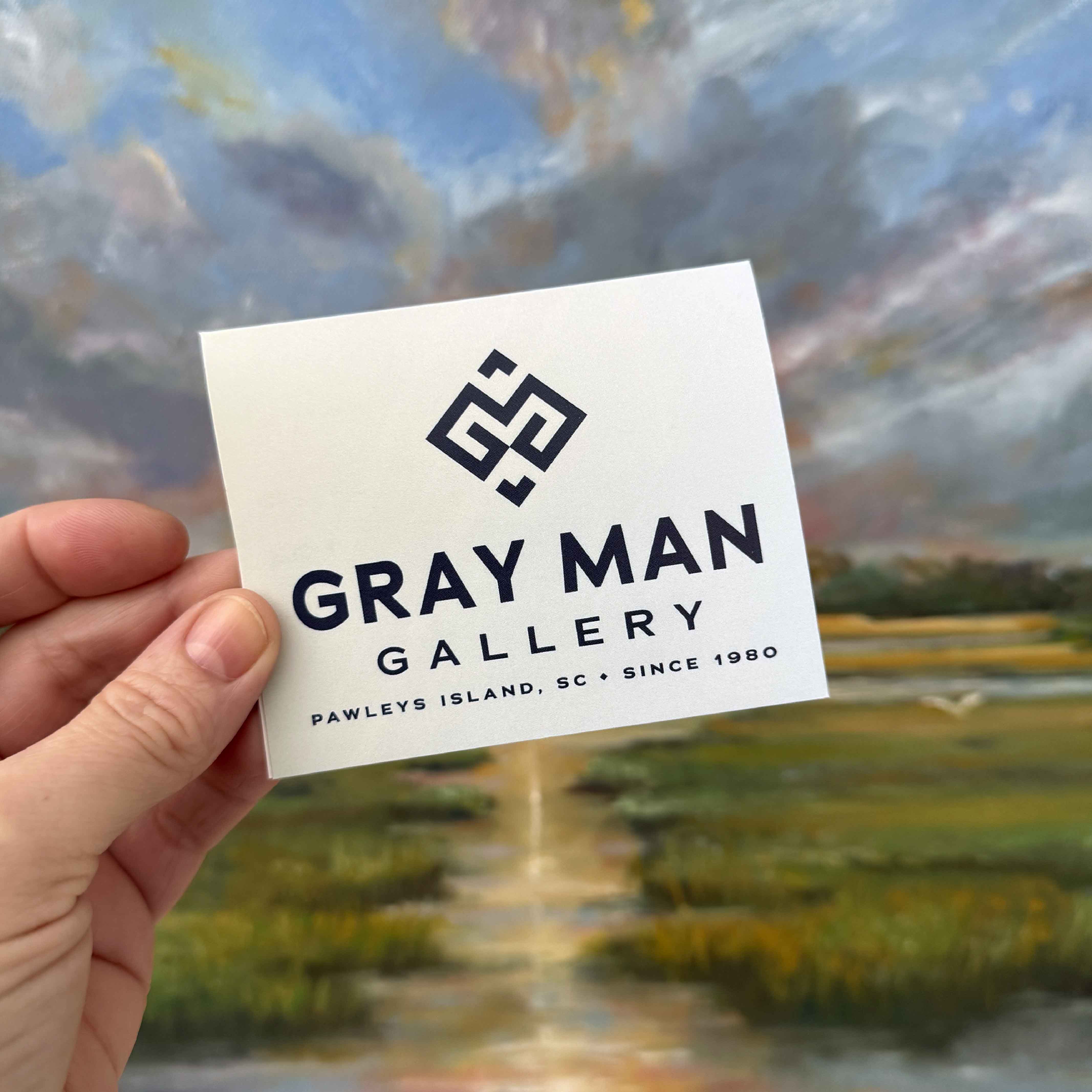 Gray Man Gallery physical gift card held in front of a Lowcountry marsh painting by local artist Betsy Boyle inside the Pawleys Island art gallery.