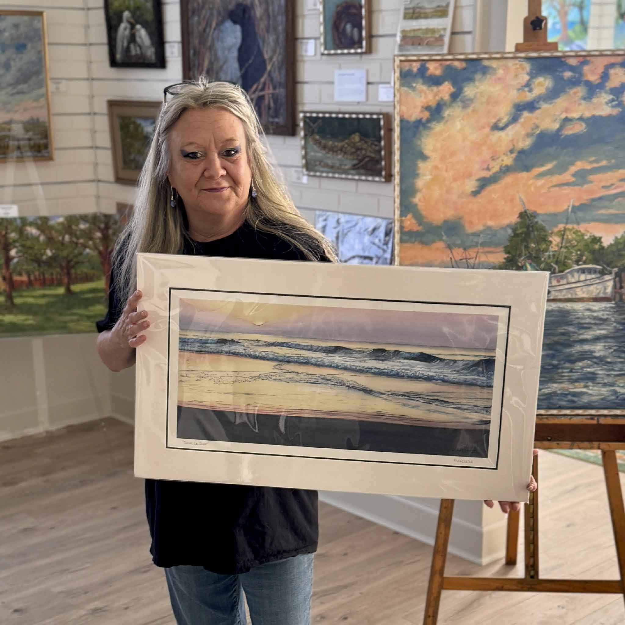 Charlotte holding a serene Lowcountry seascape print by Ron Haeberle showing rolling surf at dawn, with pink and lavender skies reflecting across wet sand in this calming coastal artwork. Shown at Gray Man Gallery.