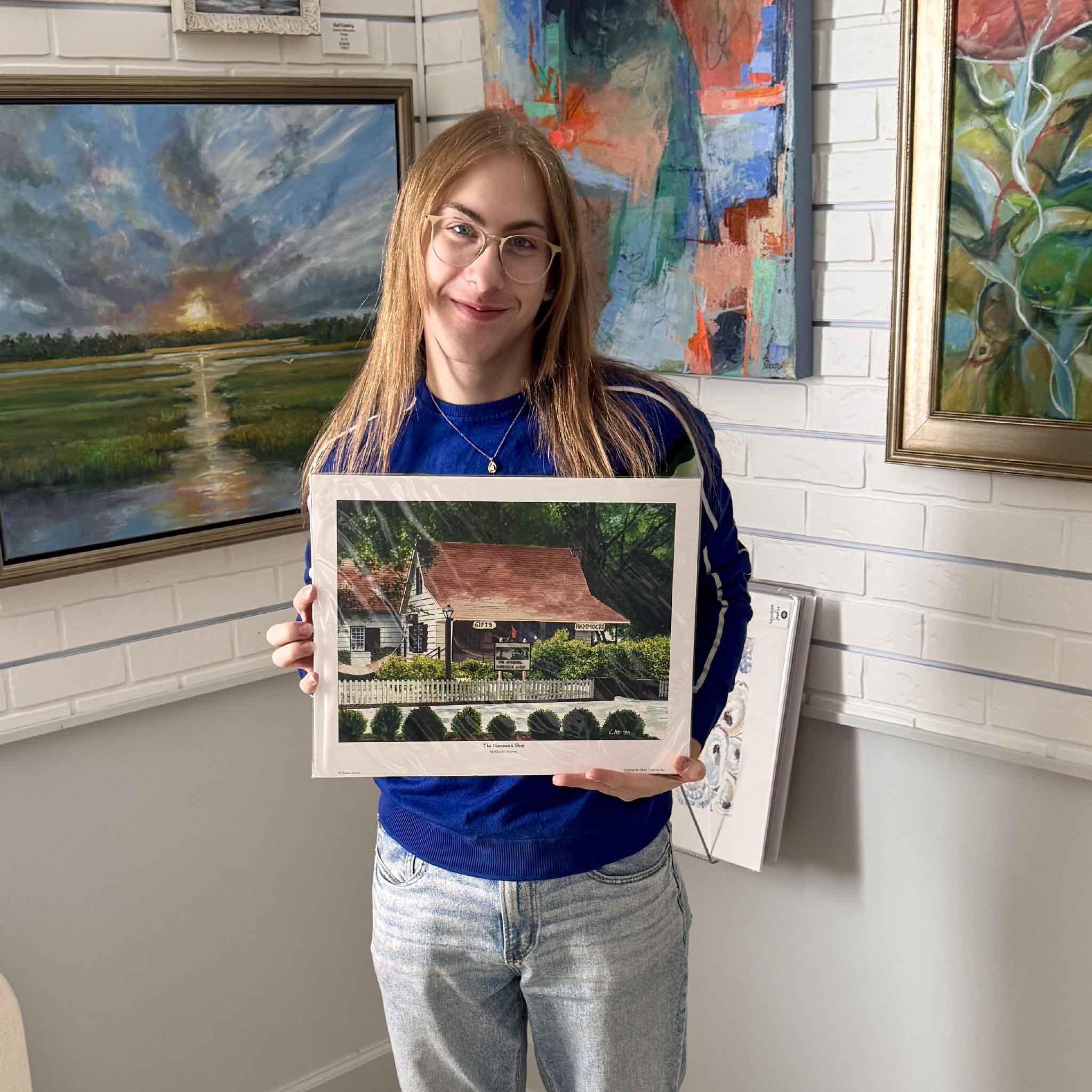 A Gray Man Gallery team member displays The Original Hammock Shop by Charles Norton in Pawleys Island, South Carolina. Featuring the landmark's famous red roof, white fence, and live oak canopy, this nostalgic Pawleys Island print fits a standard 12x16 frame, making it easy to display without custom framing.
