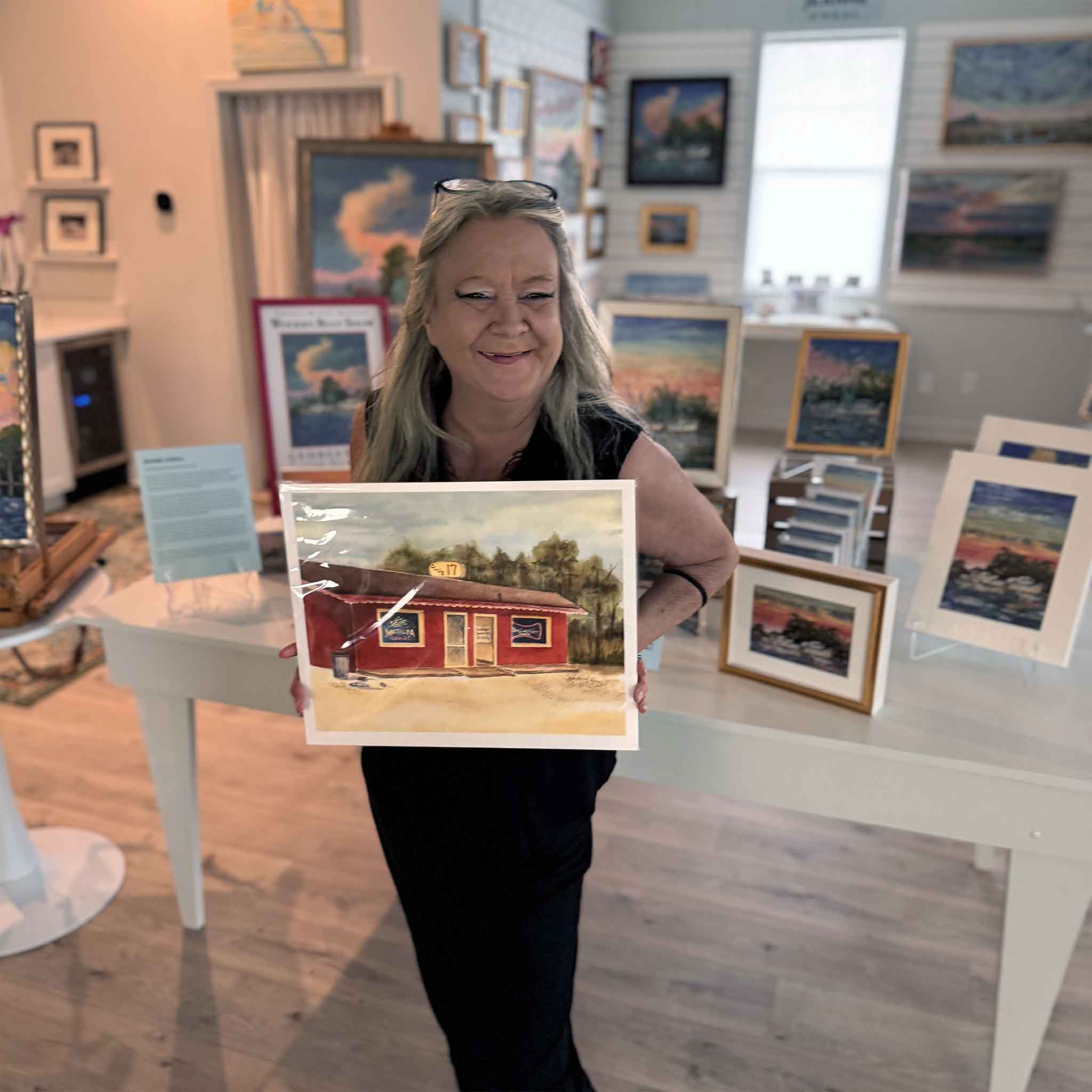 Charlotte holding Vida Miller's watercolor painting of Club 17 inside Gray Man Gallery in Pawleys Island, showcasing a nostalgic Lowcountry landmark rendered in warm tones and classic coastal style.