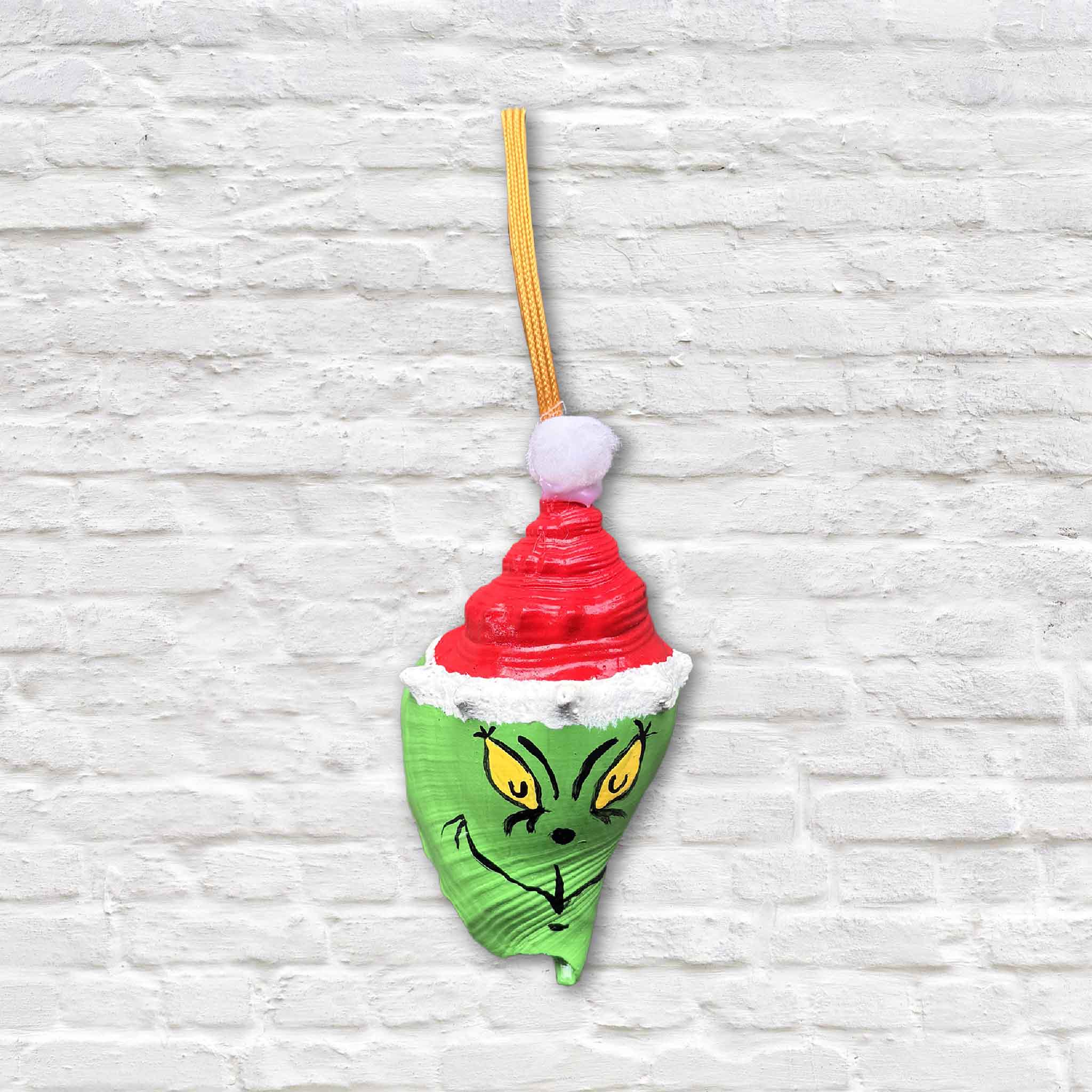 This image shows a hand-painted Grinch conch shell ornament by Tootsie Watkins, featuring a bright green Grinch face wearing a red Santa hat against a white brick background.