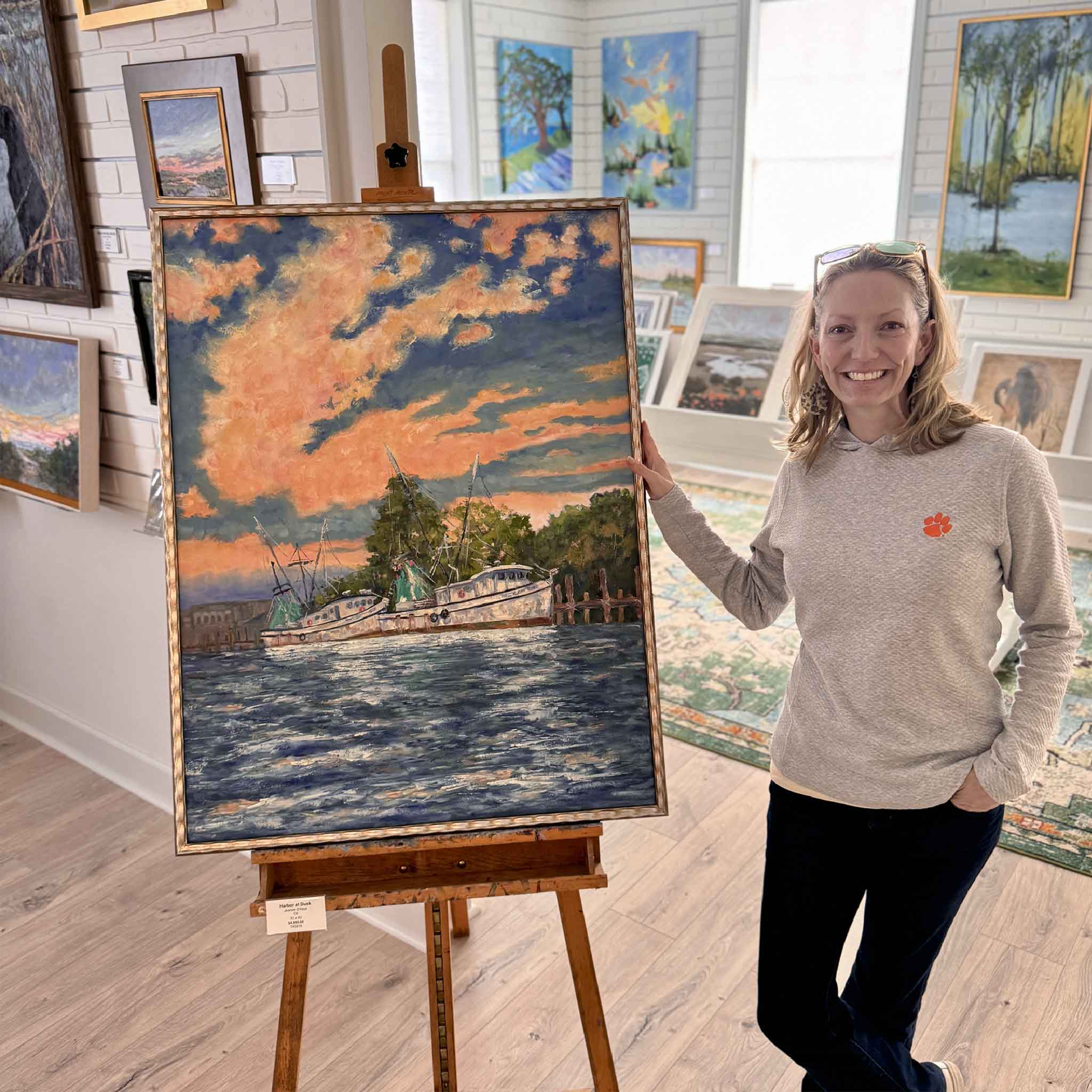Artist Jeanne O’Neal standing beside her large oil painting “Harbor at Dusk,” showing artwork scale inside Gray Man Gallery.