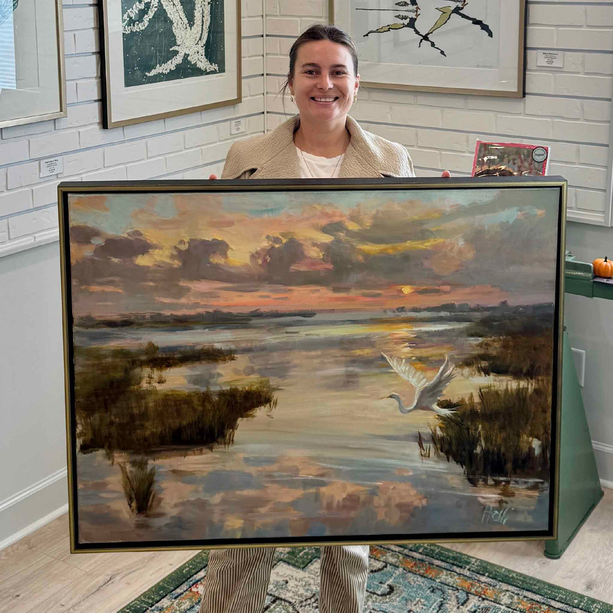 Gray Man Gallery team member holding Rising with the Dawn, a large original oil painting by local artist Helli Luck, inside Gray Man Gallery in Pawleys Island, South Carolina. The horizontal coastal landscape features a white egret flying over reflective tidal waters and golden marsh grasses beneath a pastel sunrise sky. The photograph highlights the painting's substantial scale and elegant gold floater frame.