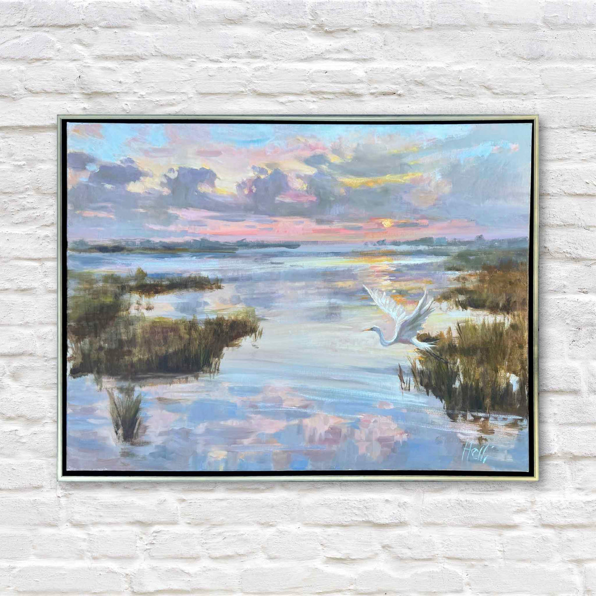 Rising with the Dawn, an original oil painting by local South Carolina artist Helli Luck, depicting a white egret taking flight above a peaceful Lowcountry tidal marsh at sunrise. Soft lavender, pink, blue, and golden clouds reflect across winding waterways surrounded by coastal marsh grasses. Presented in a slender gold floater frame against the white brick wall at Gray Man Gallery in Pawleys Island, South Carolina.