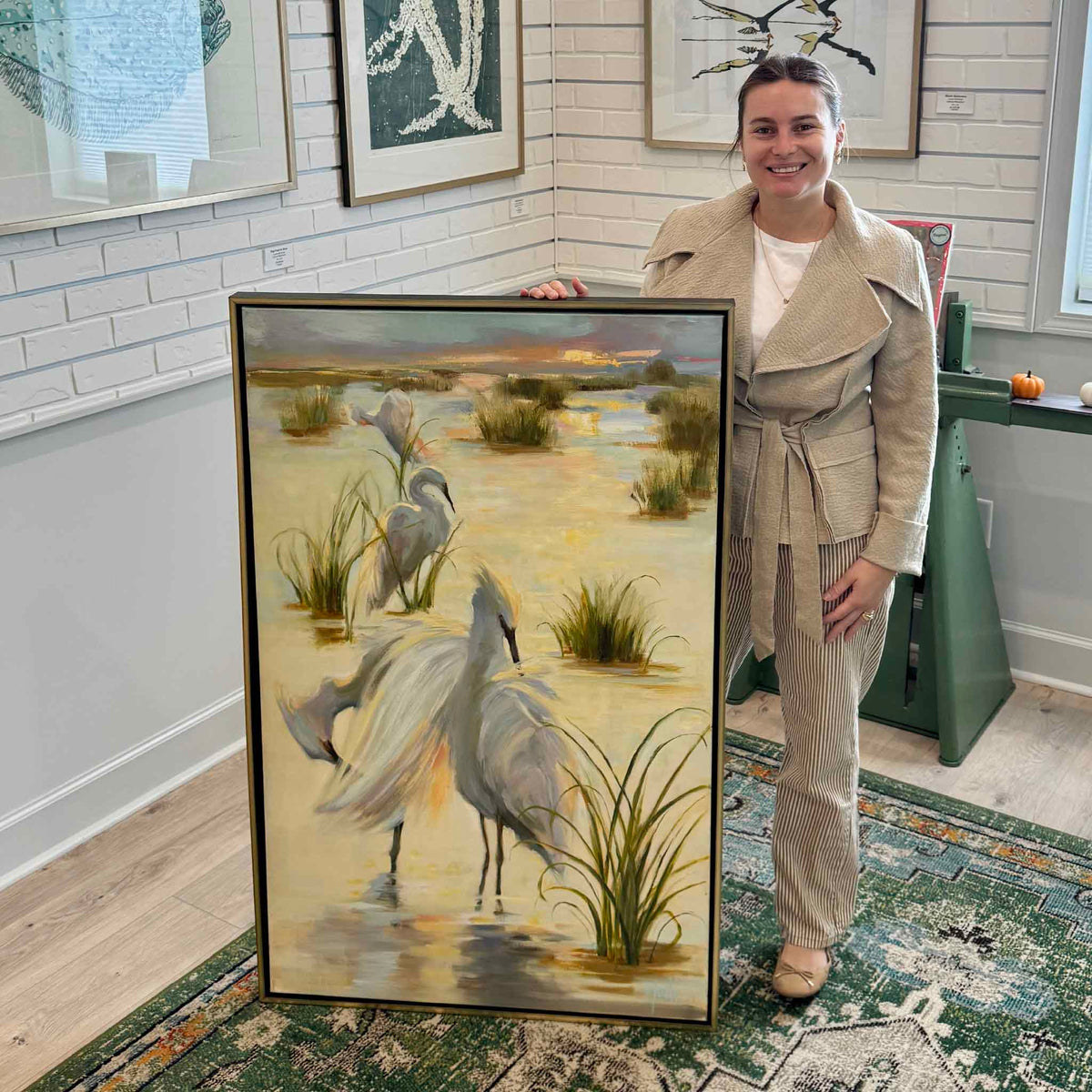 Gray Man Gallery team member standing beside Where the Water Glows, a large vertical original oil painting by local artist Helli Luck, displayed inside Gray Man Gallery in Pawleys Island, South Carolina. The framed coastal wildlife painting features three white egrets in a luminous South Carolina Lowcountry marsh and a softly glowing sunset sky. The gallery photograph illustrates the painting's impressive scale and elegant gold floater frame.