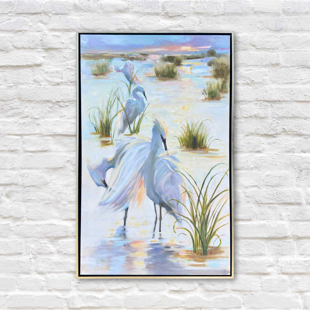 Where the Water Glows, an original oil painting by local South Carolina artist Helli Luck, featuring three elegant white egrets wading among golden-green marsh grasses in a peaceful Lowcountry tidal marsh at sunset. A large white egret spreads its wings in the foreground, its feathers painted in soft ivory, lavender, blue, and warm gold. Pastel sunset colors reflect across the water beneath a distant coastal horizon. Presented in a slender gold float frame at Gray Man Gallery in Pawleys Island.