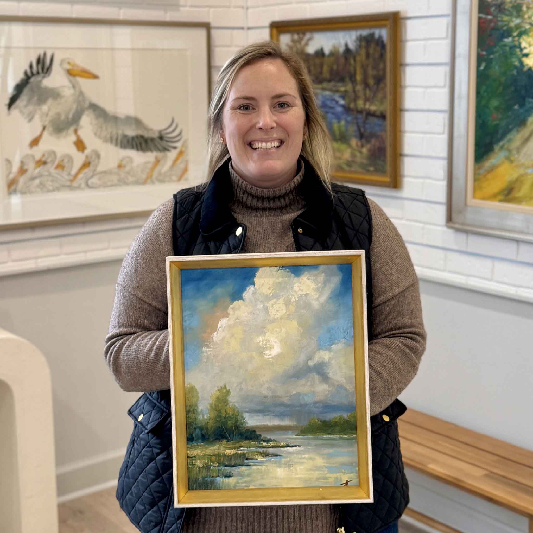 Bordy holding local artist Jamie Arnold's original oil painting “Clearing Skies” at Gray Man Gallery in Pawleys Island, South Carolina, featuring a luminous Lowcountry marsh landscape with dramatic clouds, reflective water, and custom gold frame
