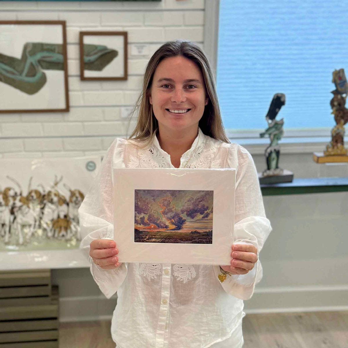Kyra holds BAM!, a matted fine art giclée print by South Carolina artist Jeanne O’Neal depicting a dramatic Lowcountry marsh sunset with storm clouds and fiery coastal light, inside Gray Man Gallery in Pawleys Island.