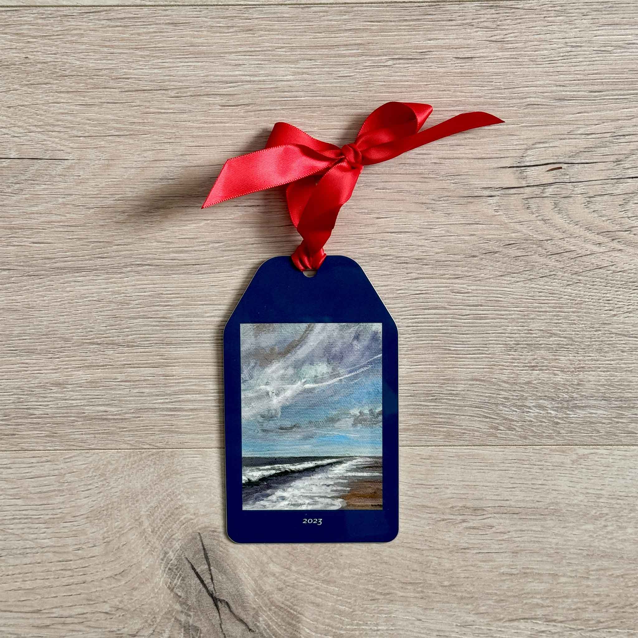 Jeanne O’Neal holiday ornament with stormy beach painting and red bow, displayed on wooden background.