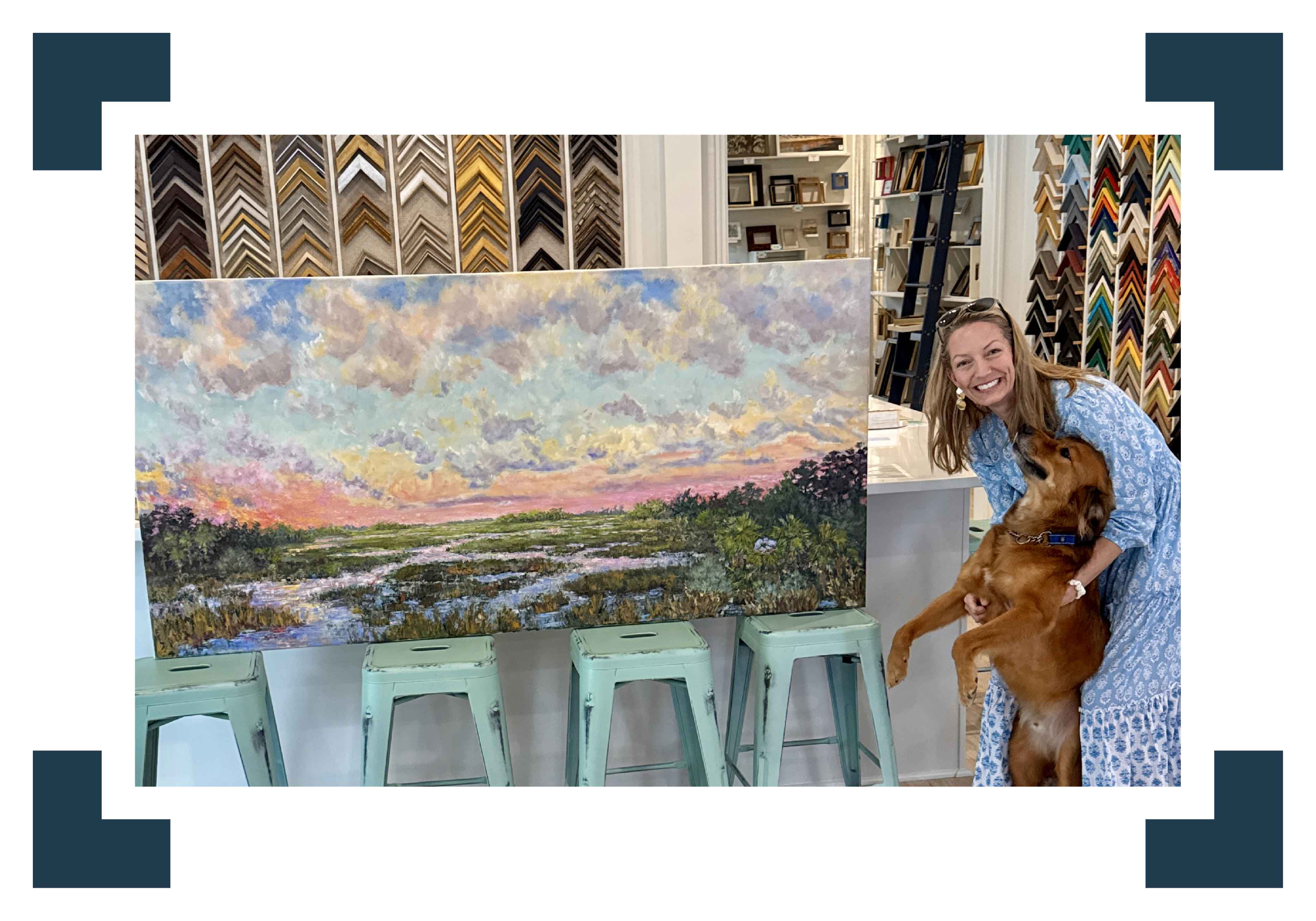 Local artist Jeanne O’Neal poses beside one of her large Lowcountry marsh landscape paintings at Gray Man Gallery in Pawleys Island while holding a golden retriever inside the gallery’s custom framing showroom.