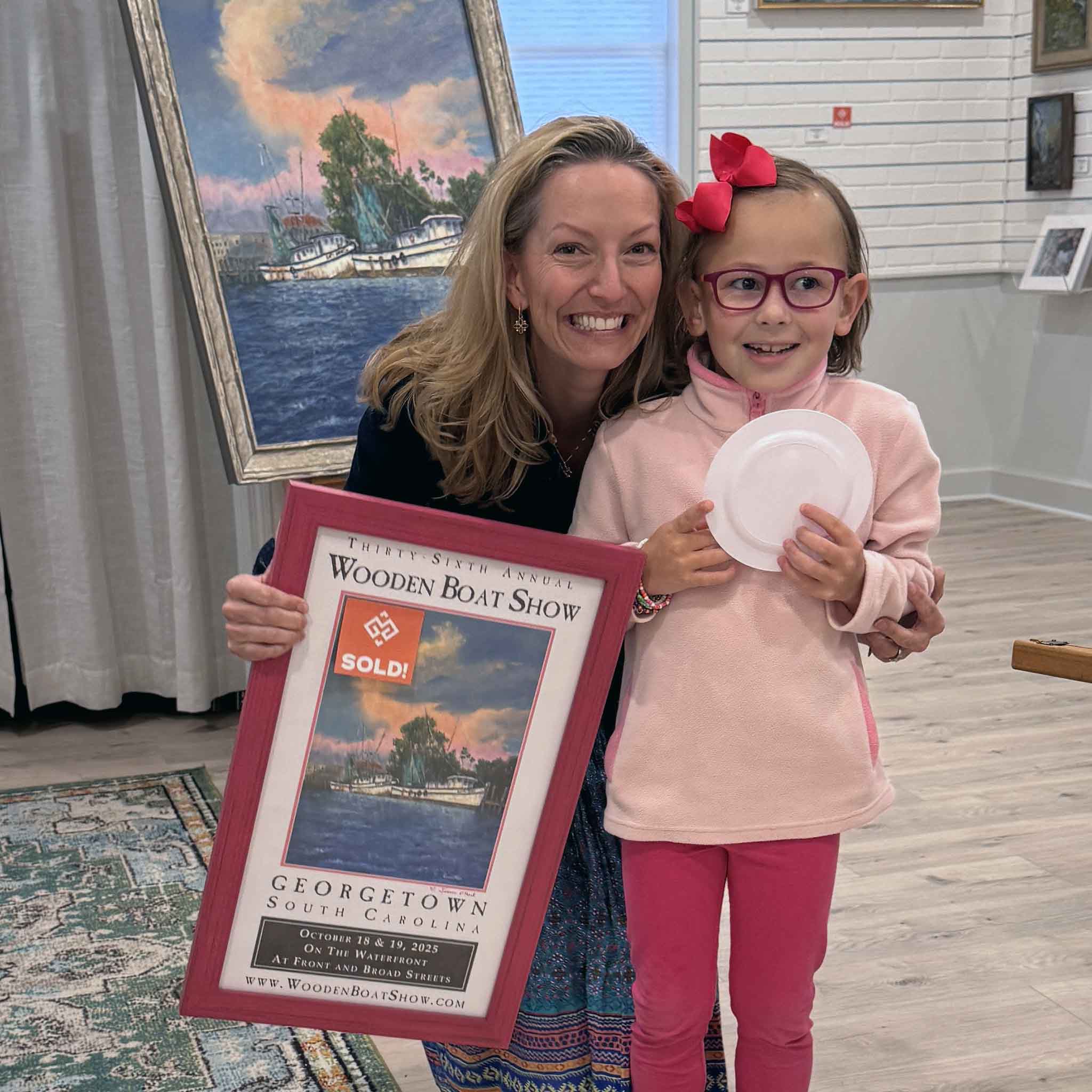 Artist Jeanne O’Neal holding her signed 2025 Georgetown Wooden Boat Show poster with a young fan at Gray Man Gallery, Georgetown SC