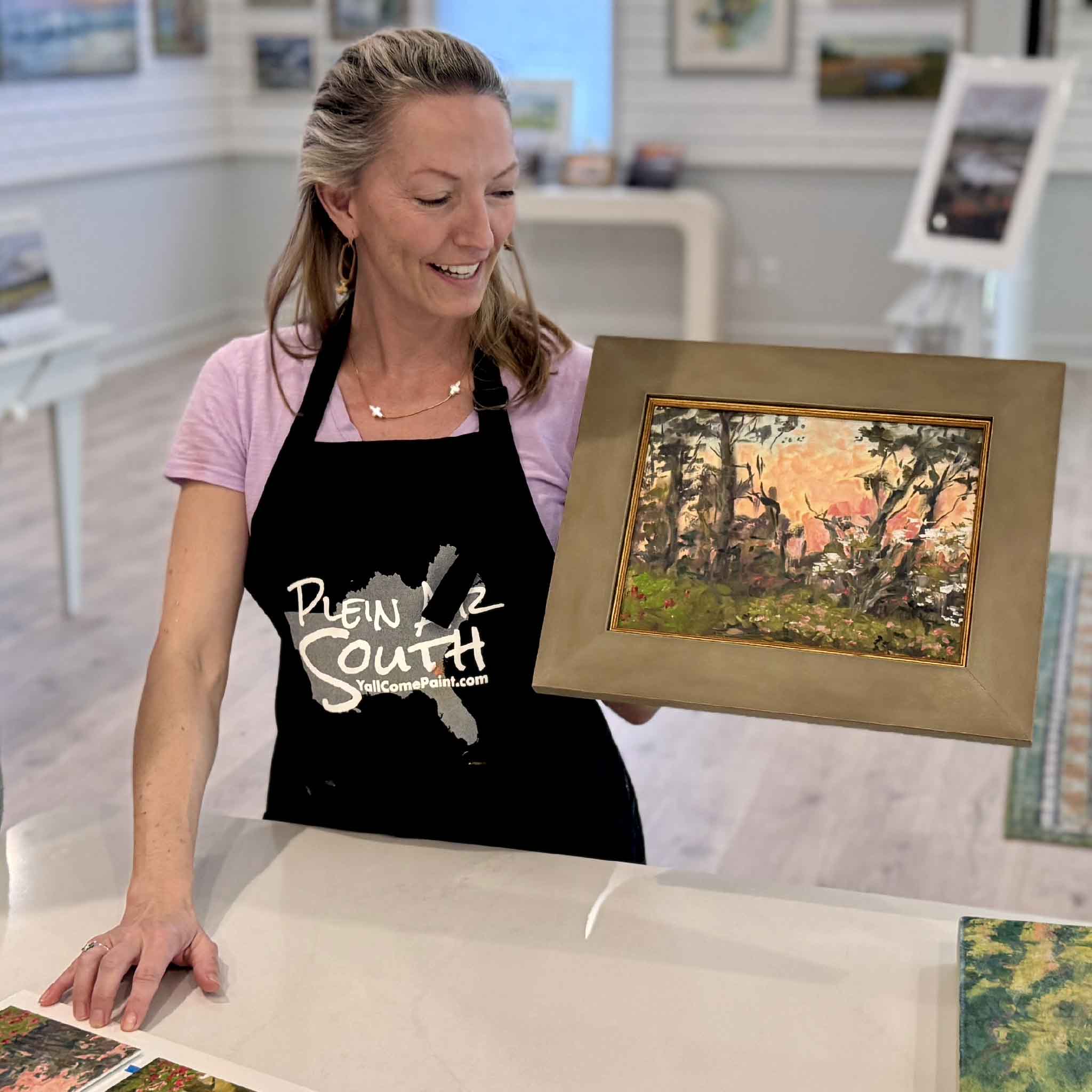 Jeanne O’Neal holding Pink Canopy original oil painting created at Brookgreen Gardens inside Gray Man Gallery Pawleys Island showing scale of framed spring floral Lowcountry landscape with pink sky