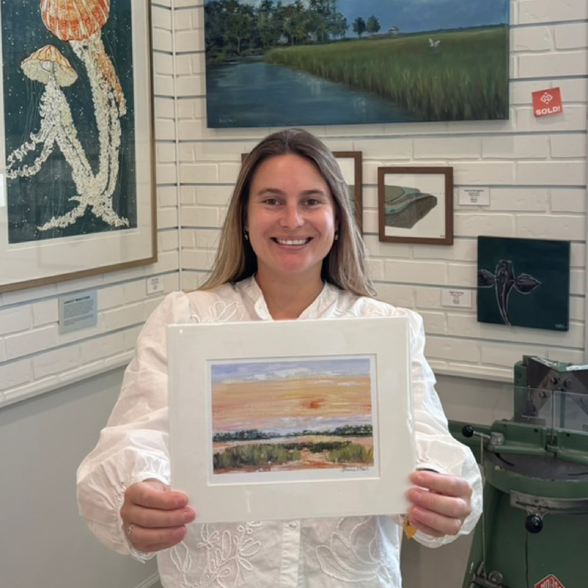 Kyra holding Jeanne O’Neal’s Low Sun fine art giclée print inside Gray Man Gallery in Pawleys Island, South Carolina, featuring a warm Lowcountry marsh sunset reproduced from the artist’s original gouache painting and professionally matted to a standard frame size.
