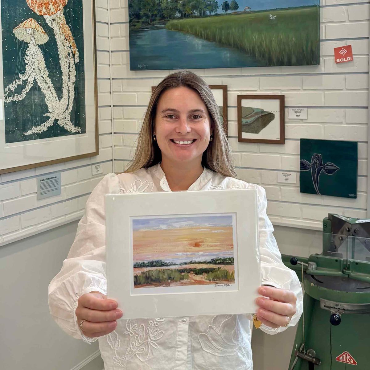 Kyra holding Jeanne O’Neal’s Low Sun fine art giclée print inside Gray Man Gallery in Pawleys Island, South Carolina, featuring a warm Lowcountry marsh sunset reproduced from the artist’s original gouache painting and professionally matted to a standard frame size.