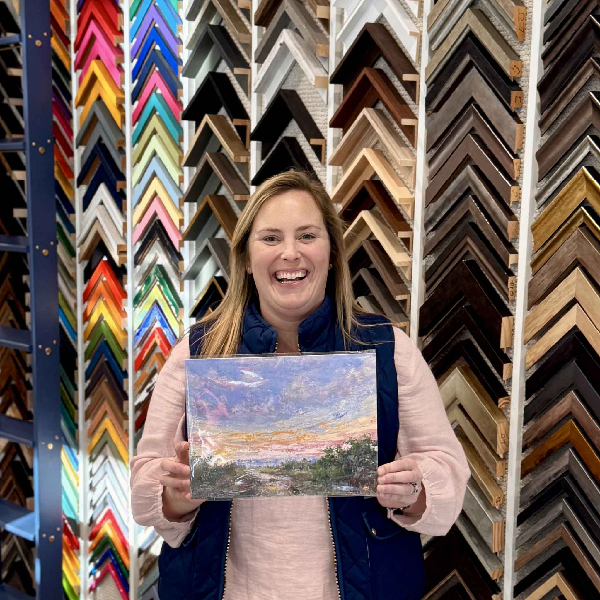 Bordy holding the 9x12 fine art giclée print of Marshlight by local South Carolina artist Jeanne O’Neal inside Gray Man Gallery in Pawleys Island, showing the vibrant Lowcountry sunset sky in coral, lavender, and gold over tidal marsh water and grasses, printed with highest quality archival inks for rich color and texture