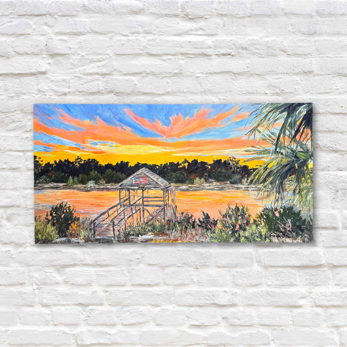 Shabby Dock, a panoramic fine art giclée print by South Carolina artist Jeanne O'Neal, depicting a weathered wooden dock and covered pier with an American flag overlooking a Lowcountry waterway at sunset. Brilliant orange, coral, yellow, and blue clouds fill the sky, reflecting across the water, while dark coastal trees, native vegetation, and sweeping palmetto fronds frame the scene. Displayed against a white brick wall at Gray Man Gallery in Pawleys Island, South Carolina.