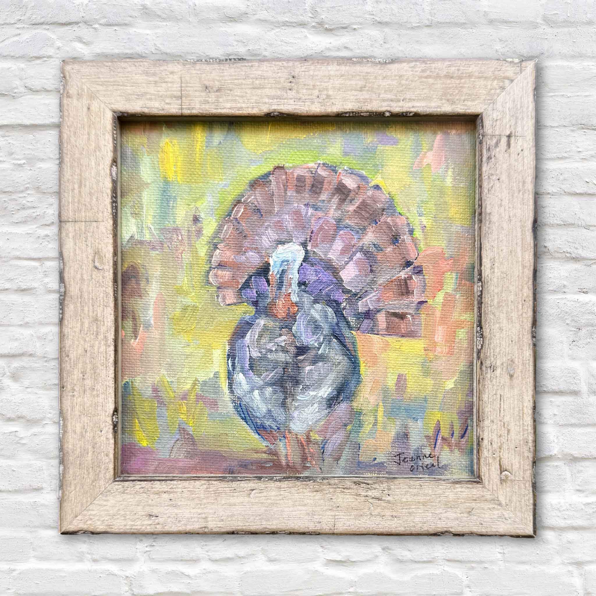Original oil painting titled Turkey Season by local artist Jeanne O'Neal featuring a colorful wild turkey standing in a field of soft greens, pinks, and golds. Framed in a rustic wood frame and photographed on a white brick wall at Gray Man Gallery in Pawleys Island, South Carolina.