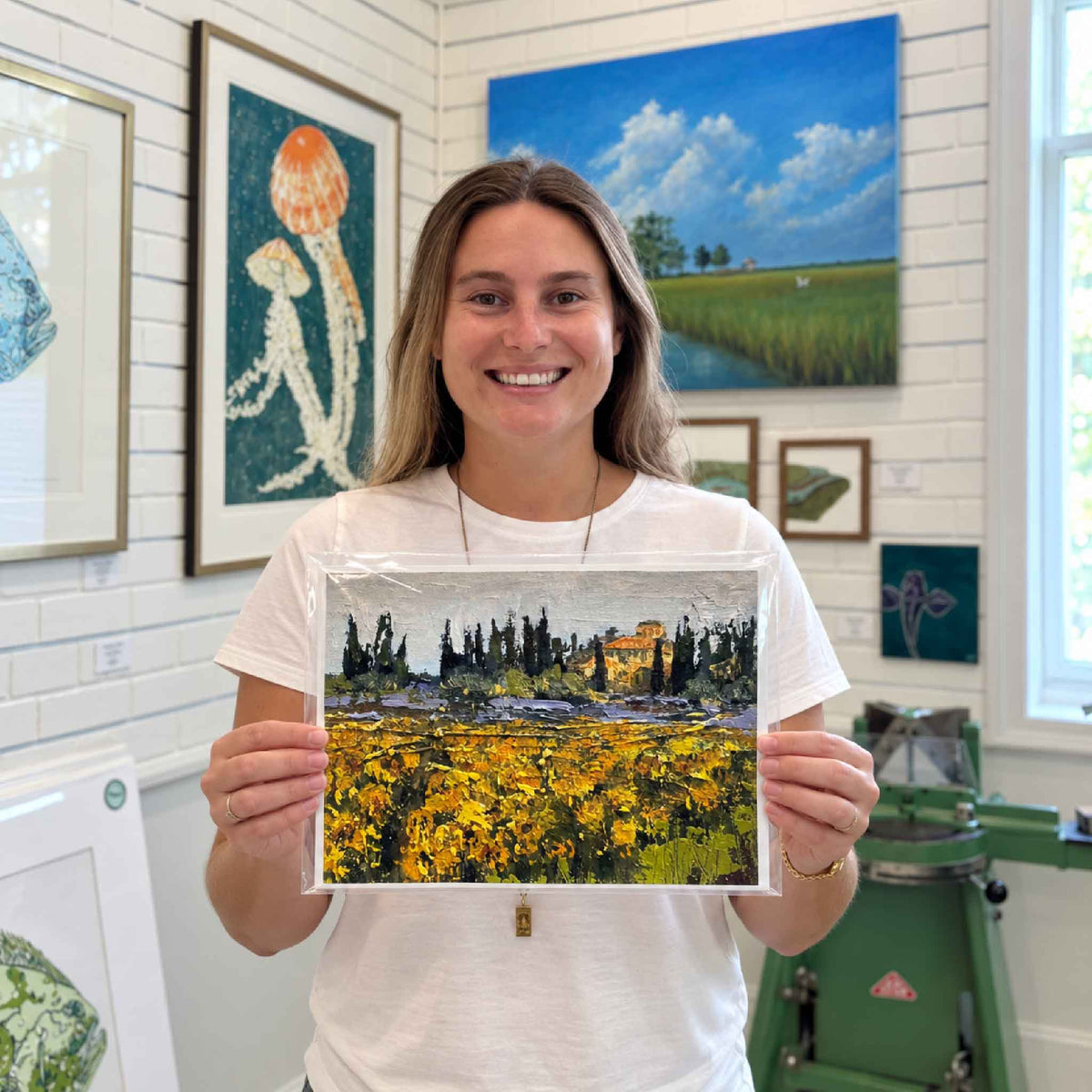 Kyra holds local artist Jeanne O’Neal’s Tuscan Spring giclée print featuring a colorful Italian landscape with golden fields and cypress trees inside Gray Man Gallery in Pawleys Island, South Carolina