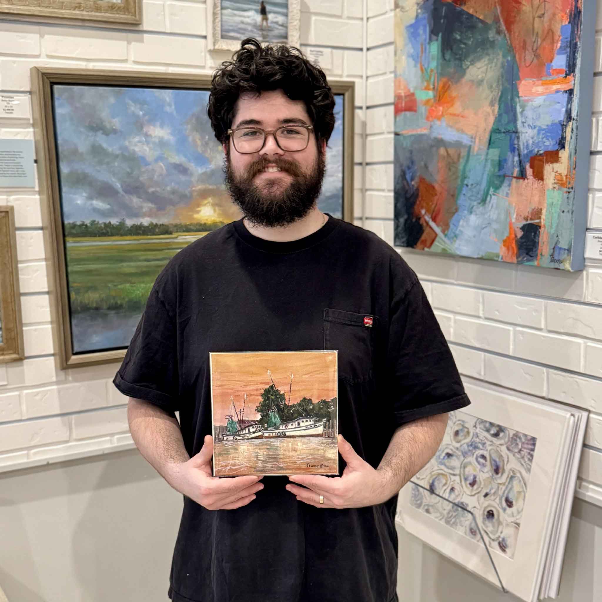 Gallery team member holding the Harbor Glow 8x8 coastal print by Lowcountry artist Jeanne O’Neal inside Gray Man Gallery, with framed paintings in the background.