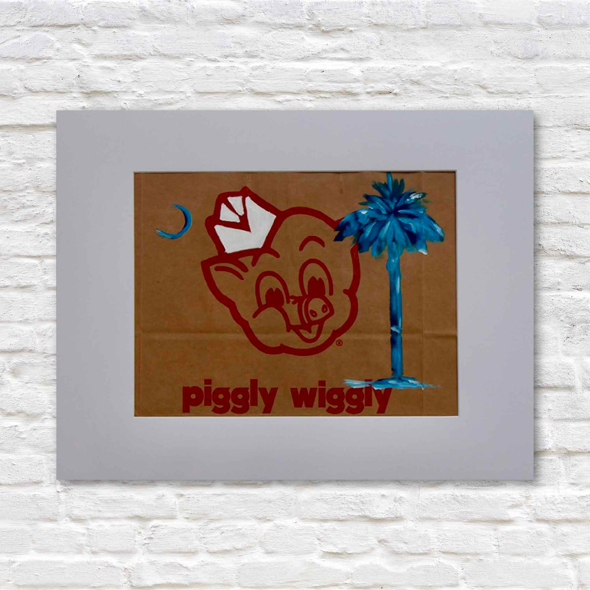 Carolina on My Mind, an original acrylic painting by local artist Jenny Reynolds featuring the blue palmetto tree and crescent moon of the South Carolina state flag painted on an authentic brown Piggly Wiggly grocery bag. The iconic red pig mascot and lettering remain visible beneath the artwork, presented in a wide light gray mat at Gray Man Gallery in Pawleys Island, South Carolina.